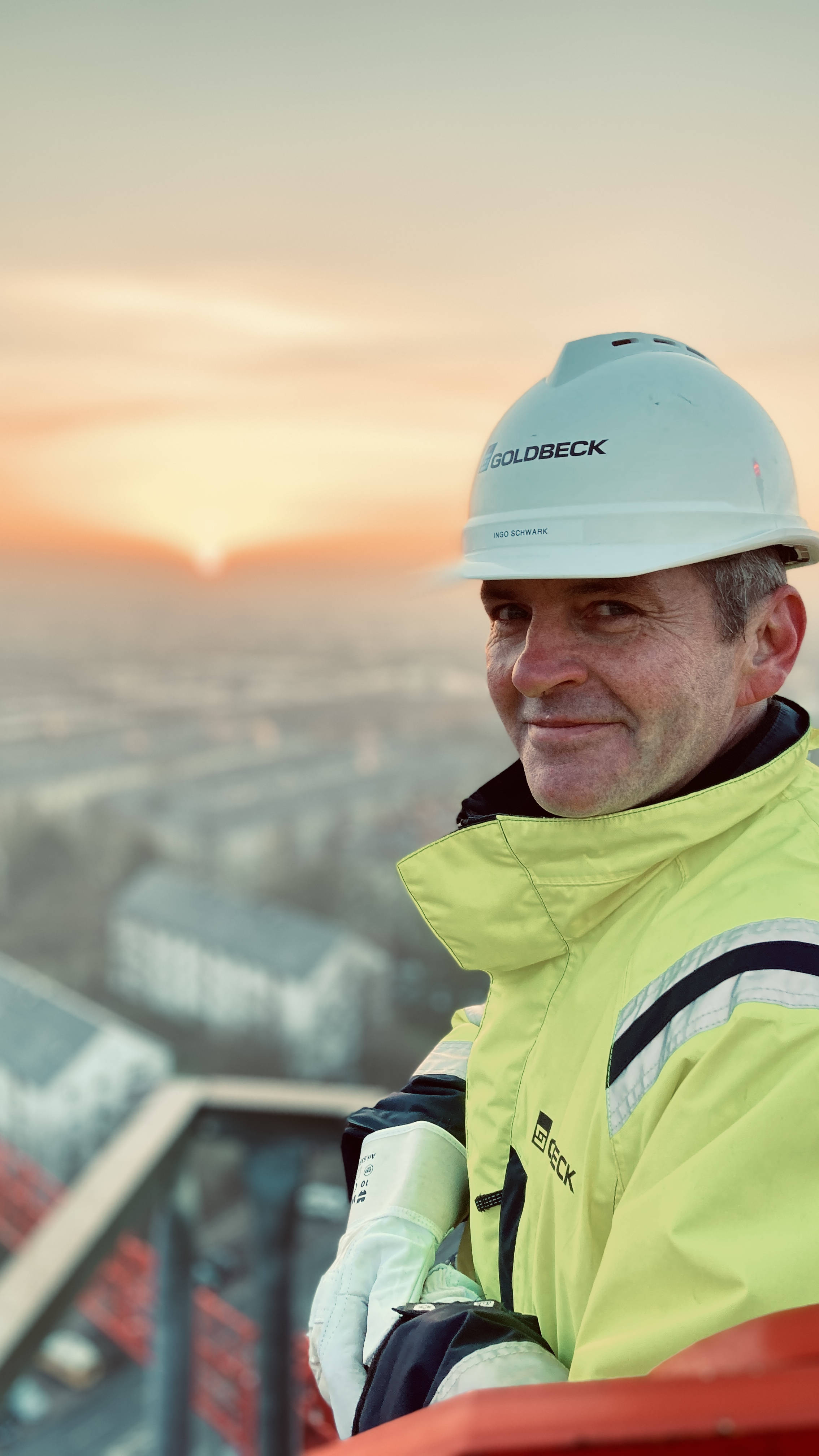 Bauarbeiter in Schutzkleidung und Helm lächelt von einer Baustelle bei Sonnenuntergang, Stadt im Hintergrund.