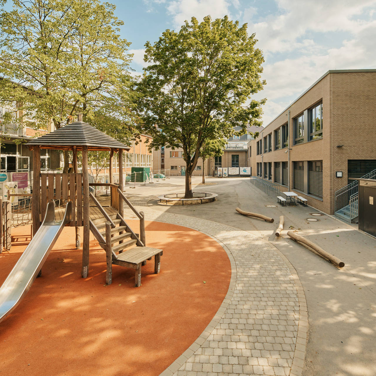 Spielplatz mit Rutsche und Klettergerüst auf dem Schulgelände, umgeben von Bäumen und modernen Gebäuden im Hintergrund.
