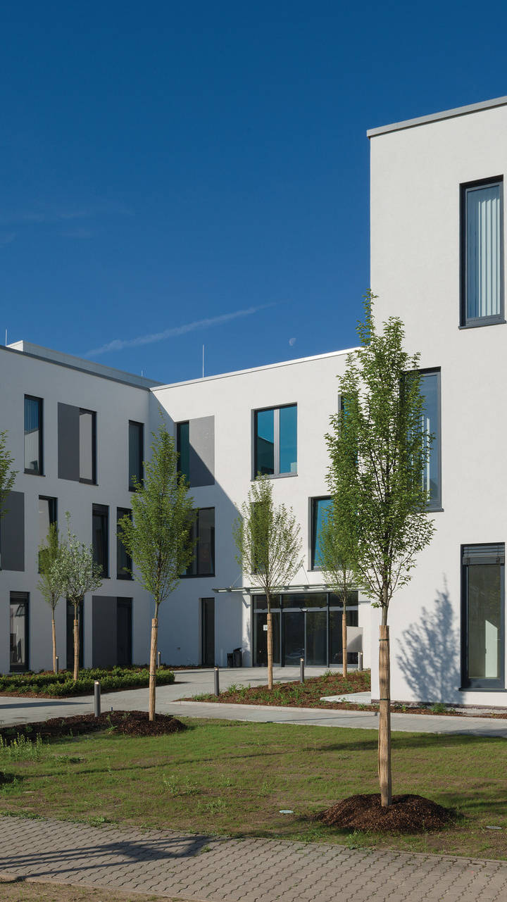 Modernes Bürogebäude mit weißen Fassaden, großen Fenstern und neu angelegten Bäumen im Vordergrund unter klarem, blauem Himmel.