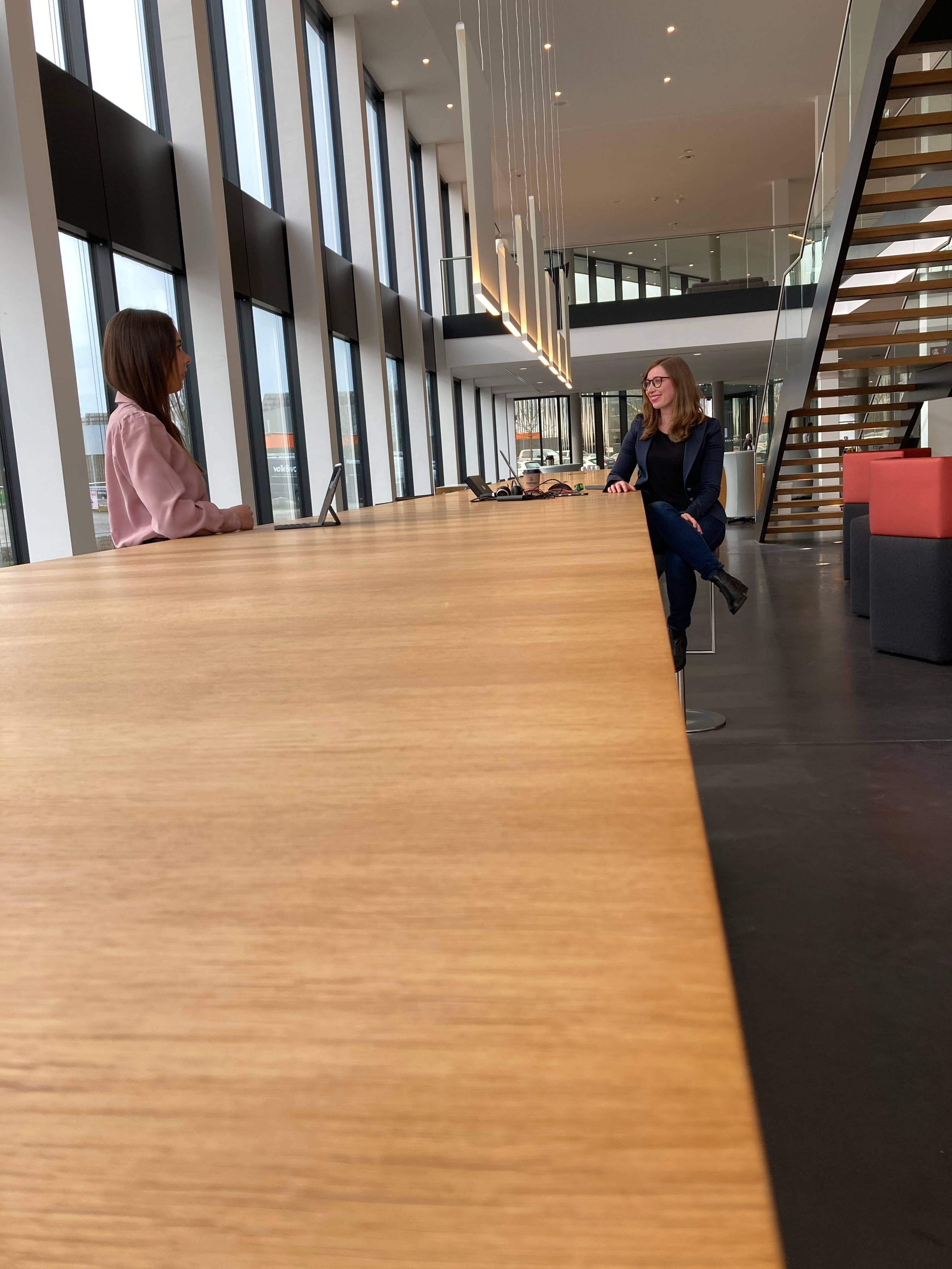 Zwei Personen sitzen in einem modernen Bürogebäude an einem langen Holztisch, großes Fenster im Hintergrund.
