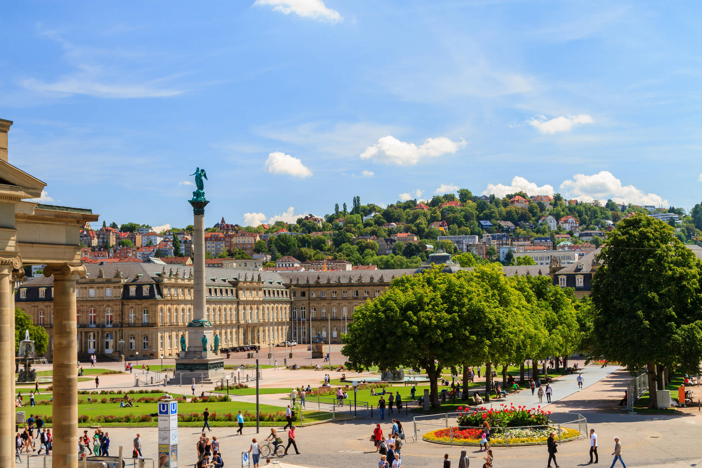 Stuttgarter Schlossplatz an einem sonnigen Tag, mit belebtem Platz, Bäumen und umliegender Architektur im Hintergrund.