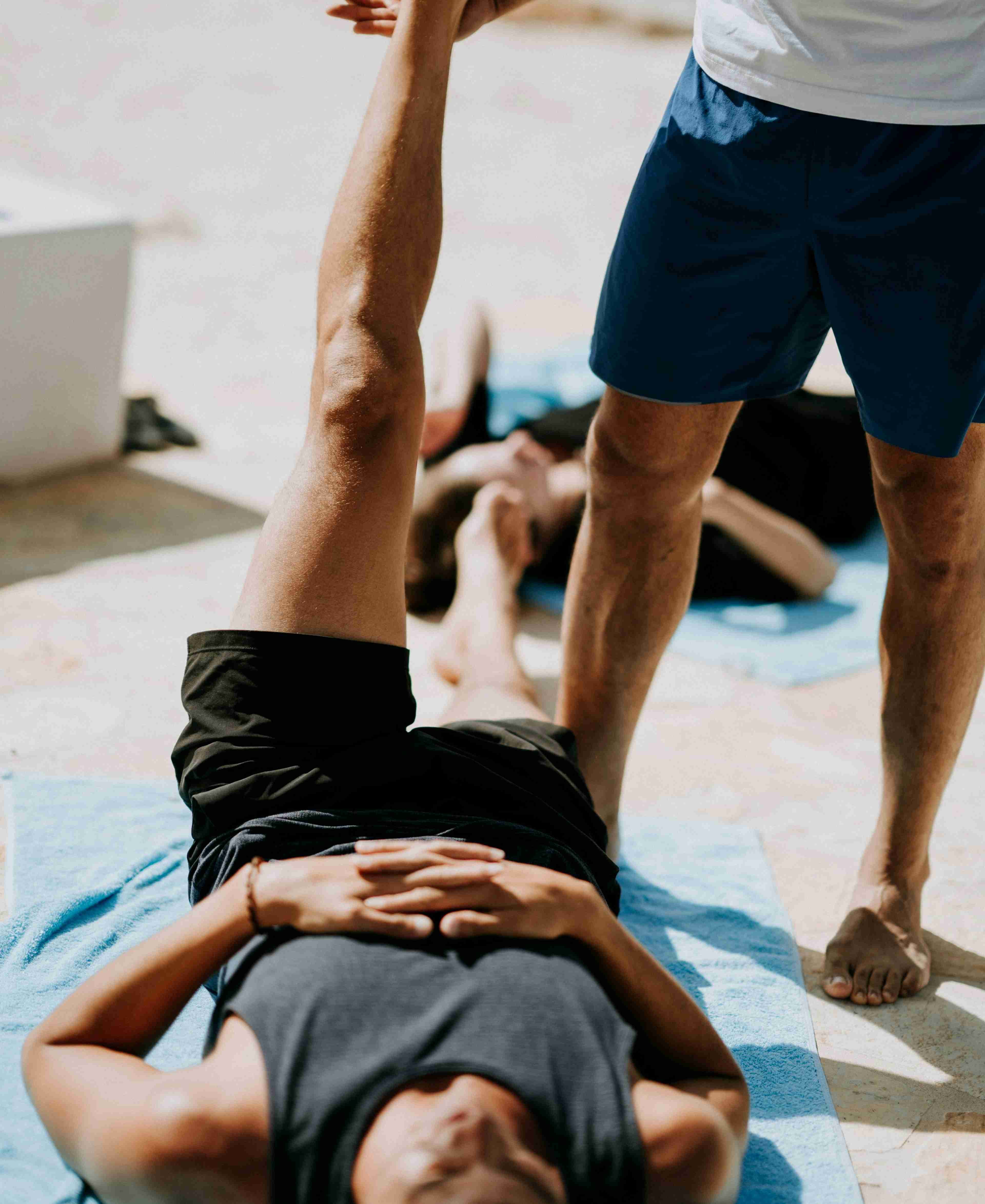 Man in a dark grey tank top lying on the floor near a man stretching his leg during a breathing exercise