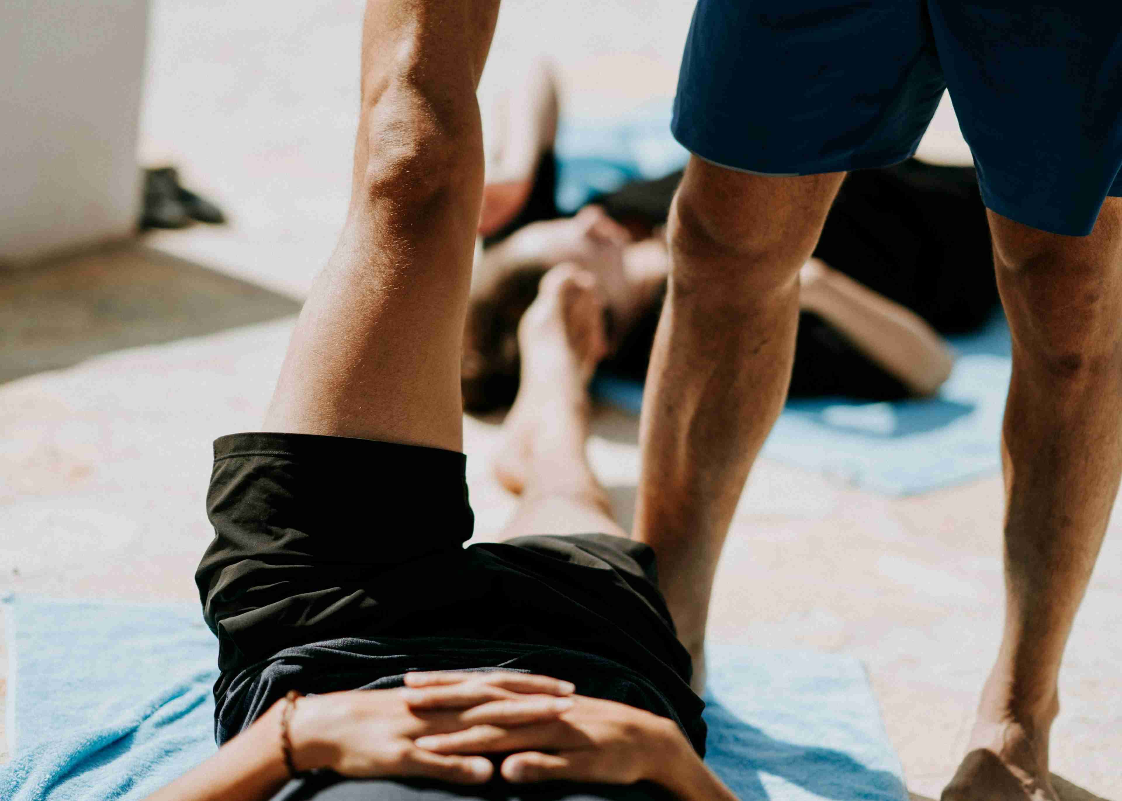 Man in a dark grey tank top lying on the floor near a man stretching his leg during a breathing exercise