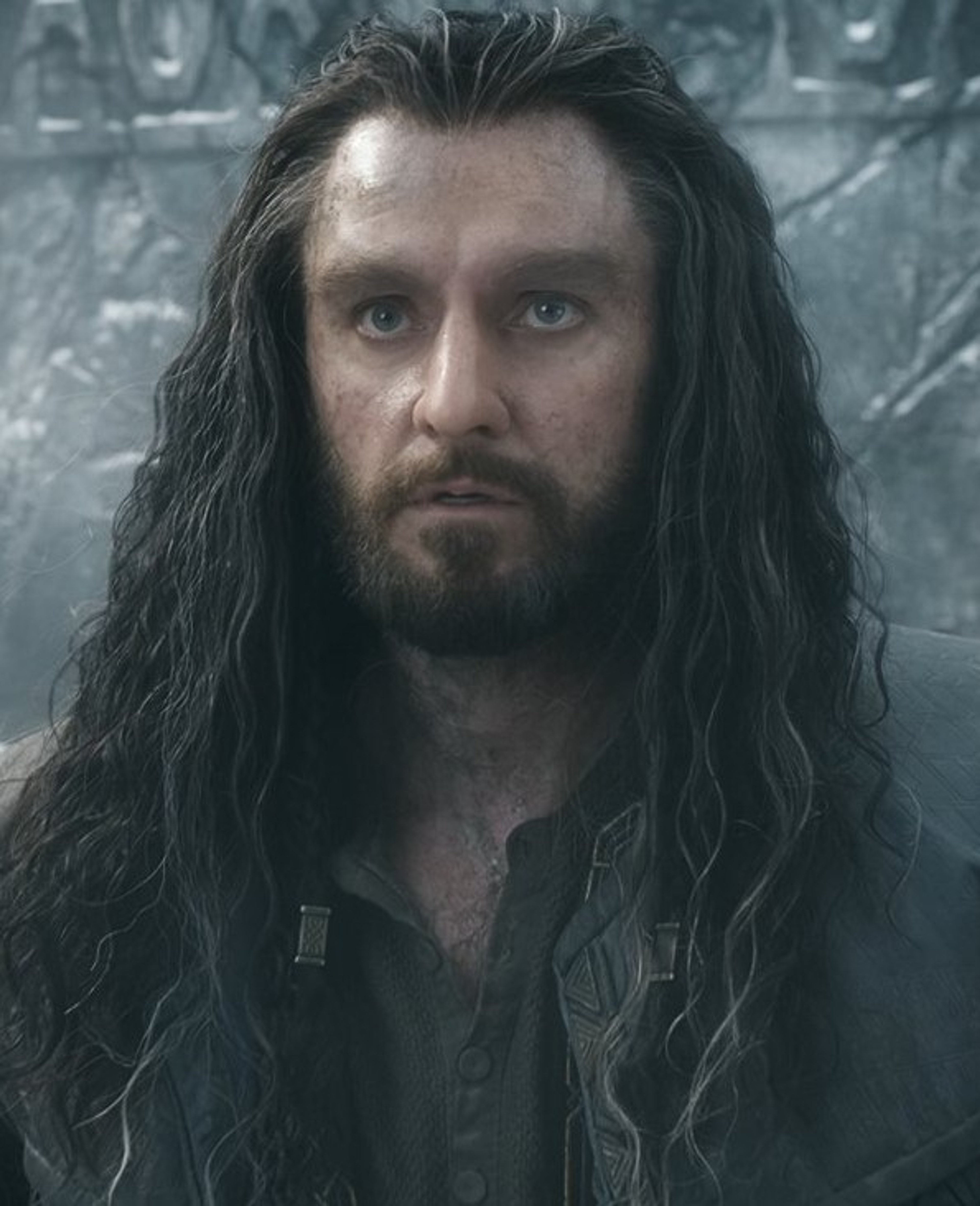 A bearded man with long dark hair in a fur-lined coat, standing in a snowy, mountainous environment.
