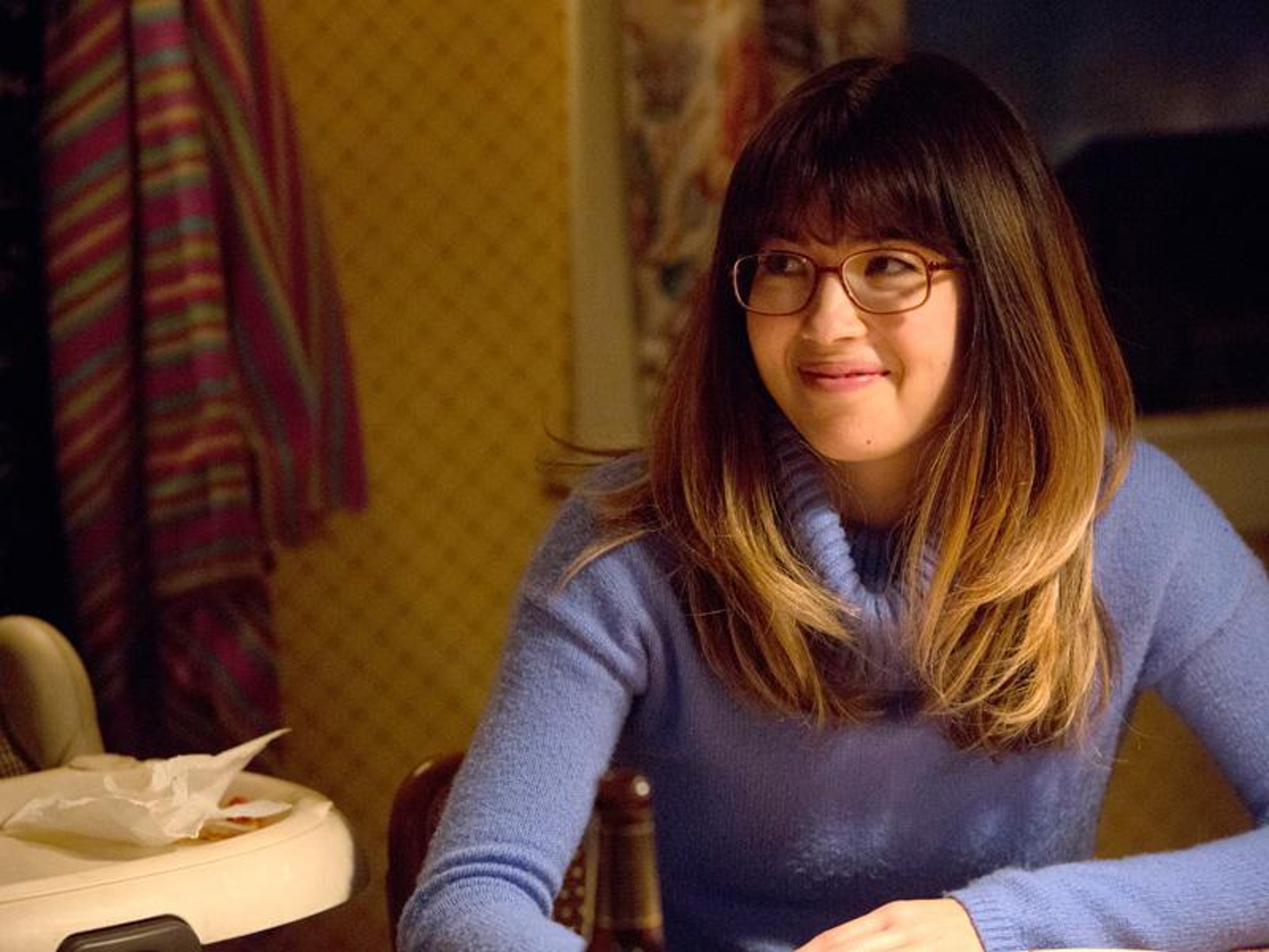 A young woman with long brown hair and glasses, wearing a blue sweater and sitting at a desk or table.