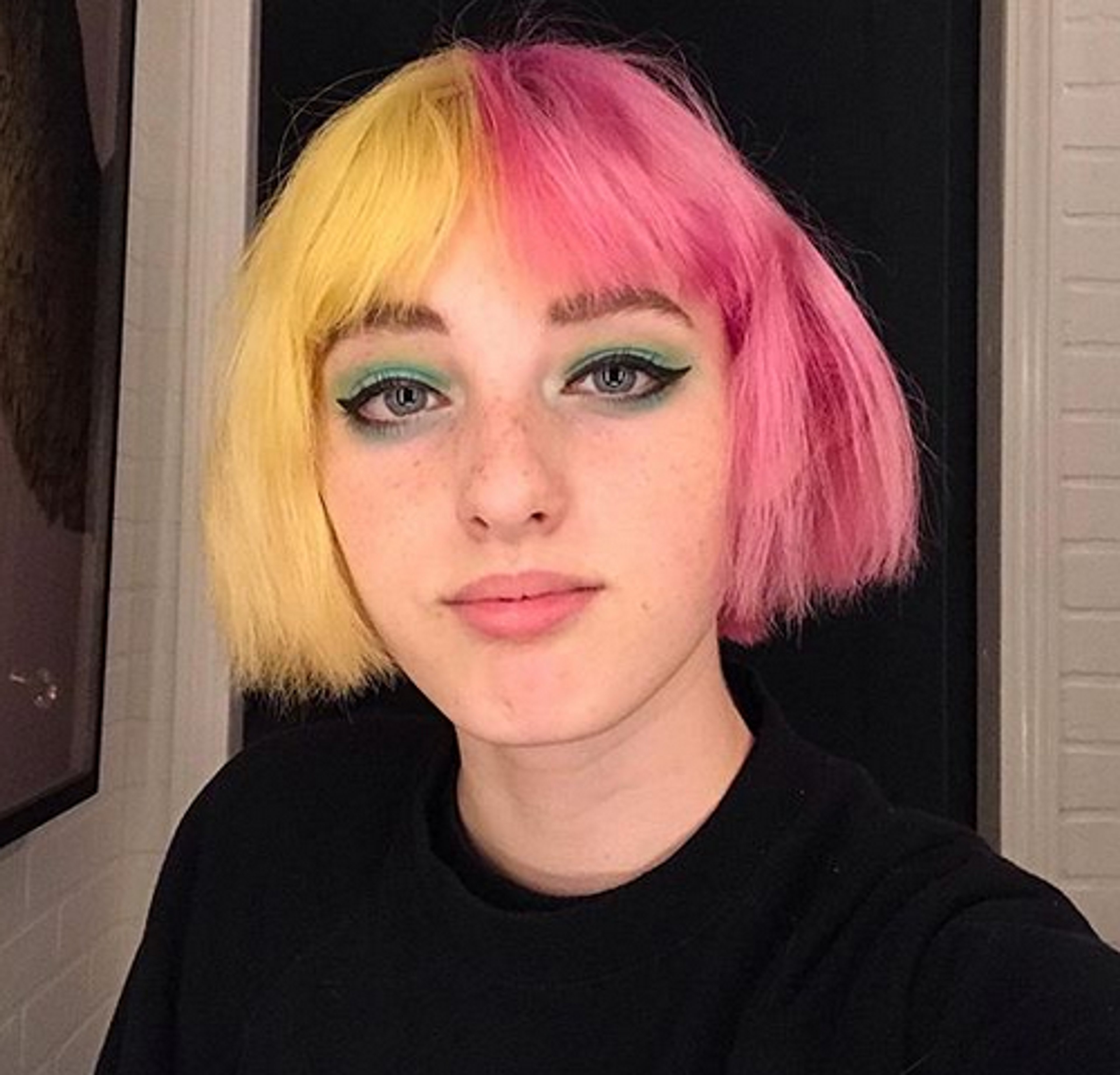 A young woman with colorful hair and green eyes, wearing a black shirt and posing for a selfie.