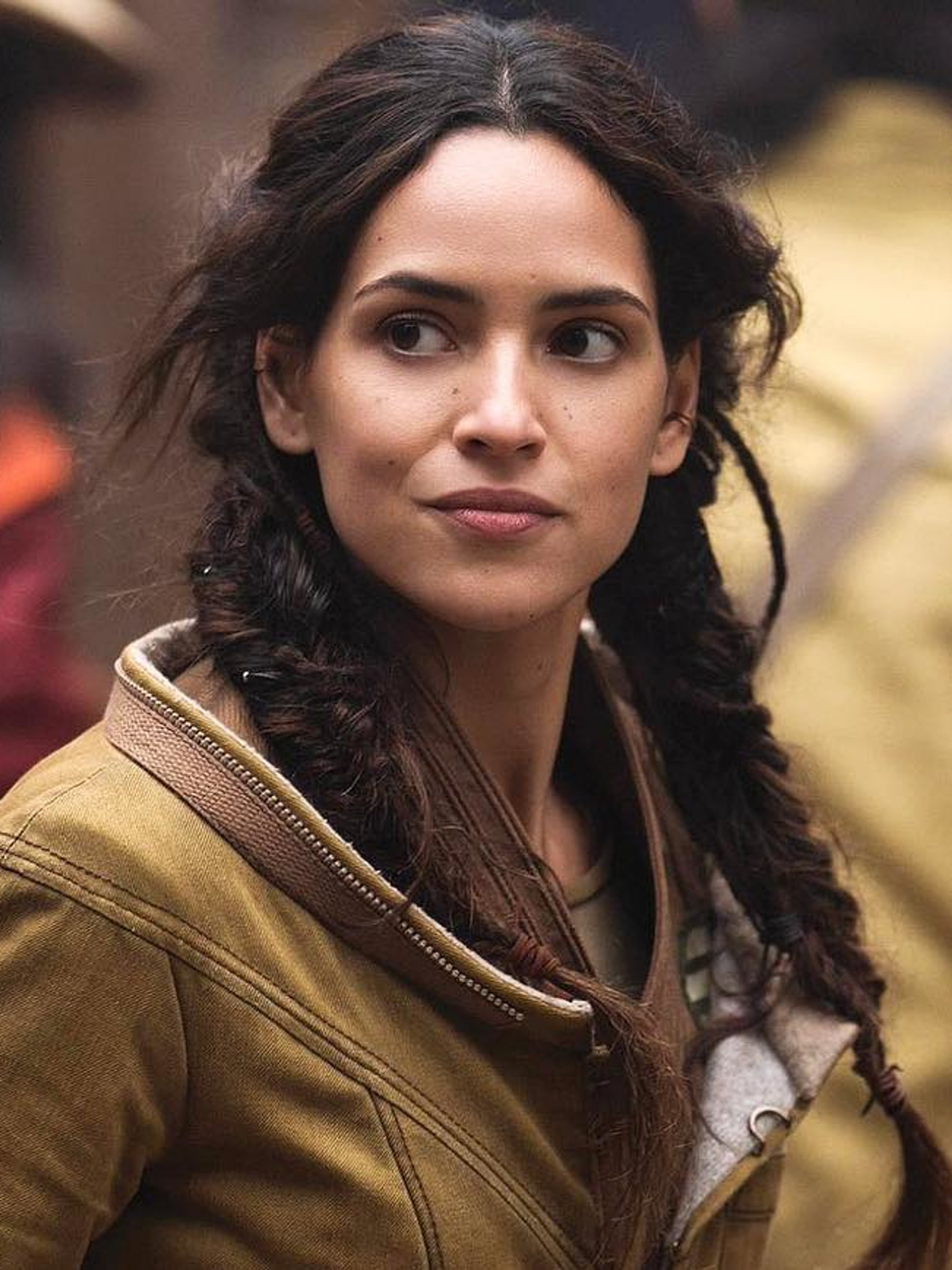 A woman with braided brown-black hair wearing a tan jacket, with a serious expression on her face.