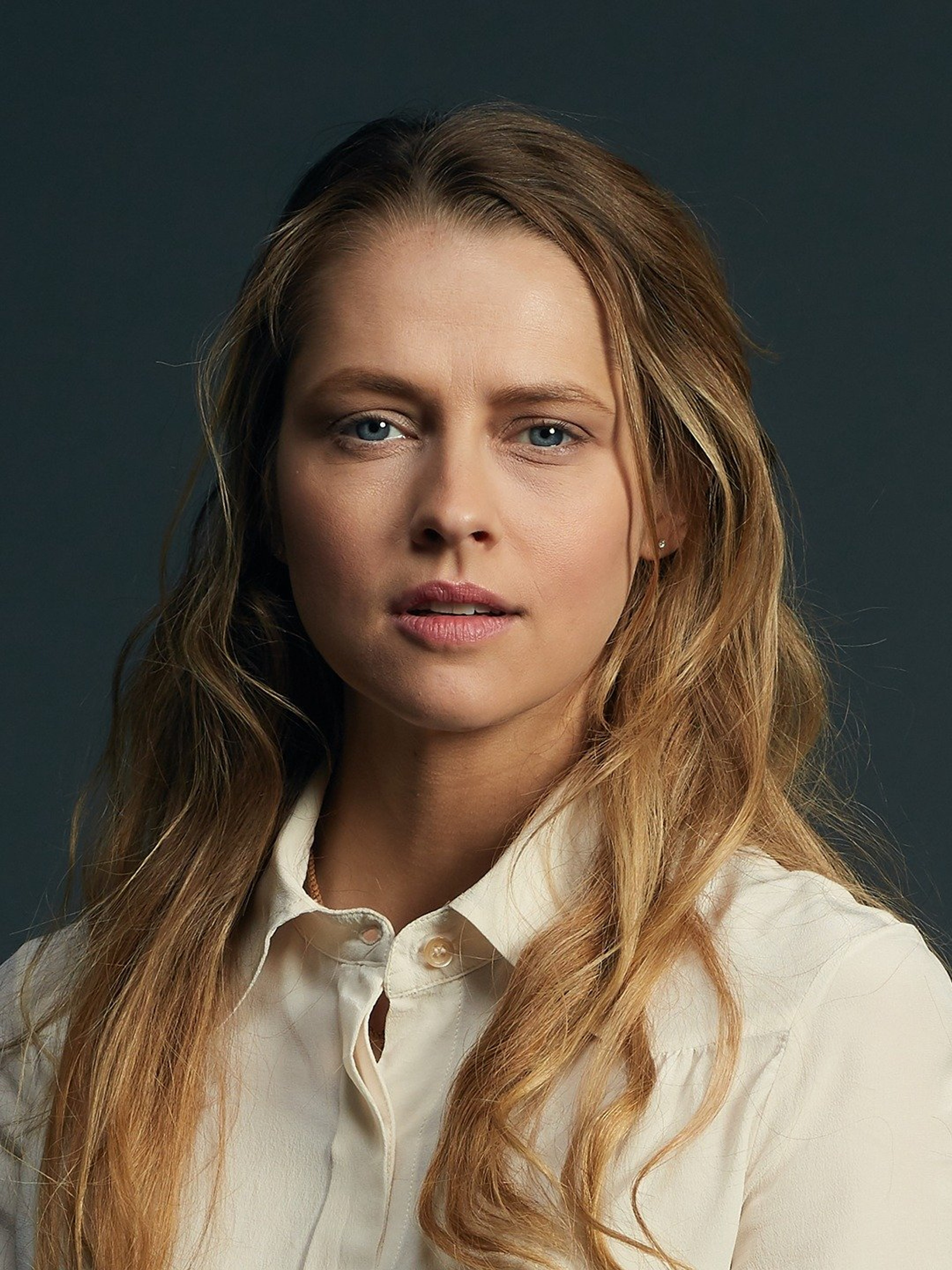 A woman with long blonde hair and blue eyes wearing a white blouse, looking thoughtful.