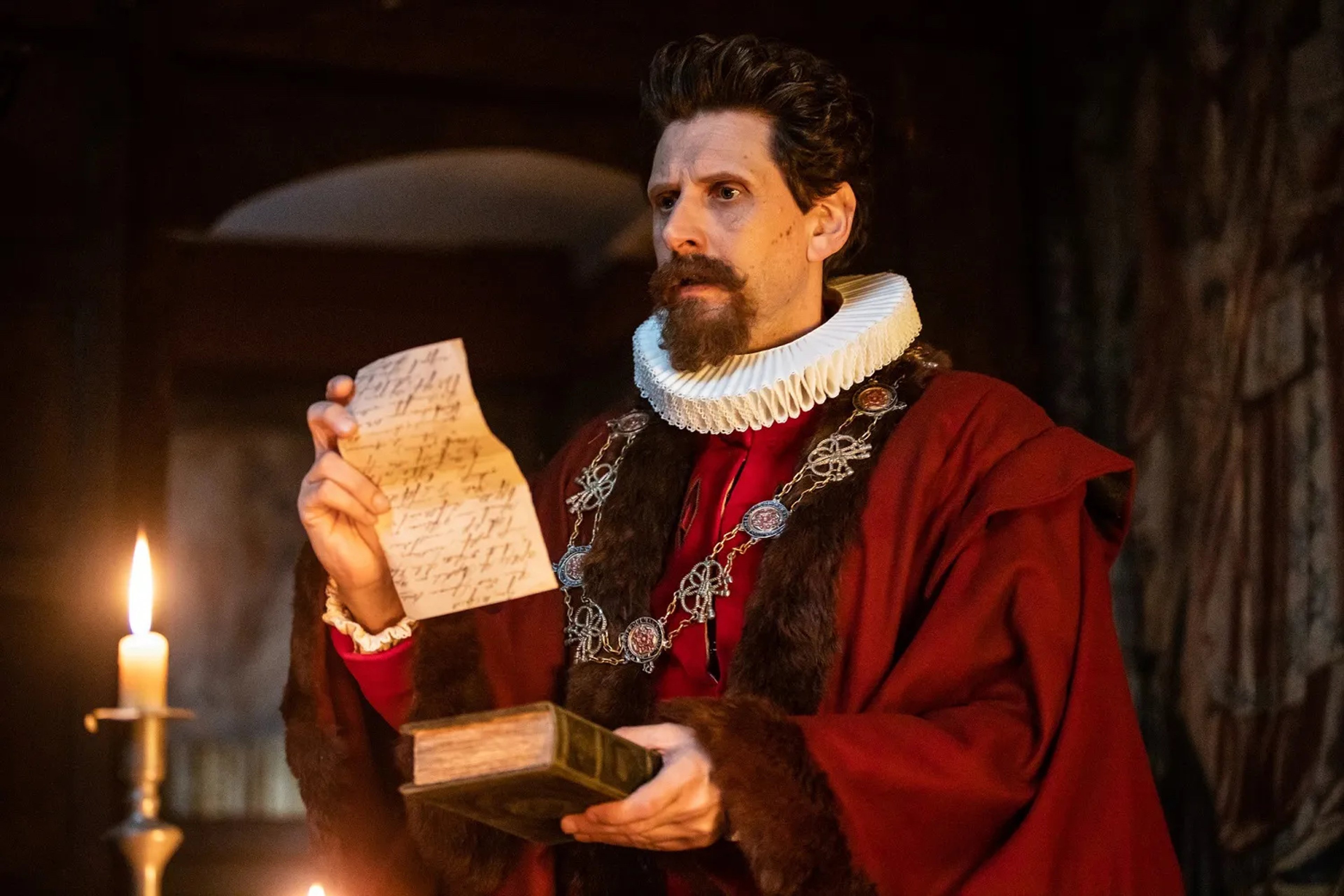 A man in a red robe holding an open book in a dimly lit room