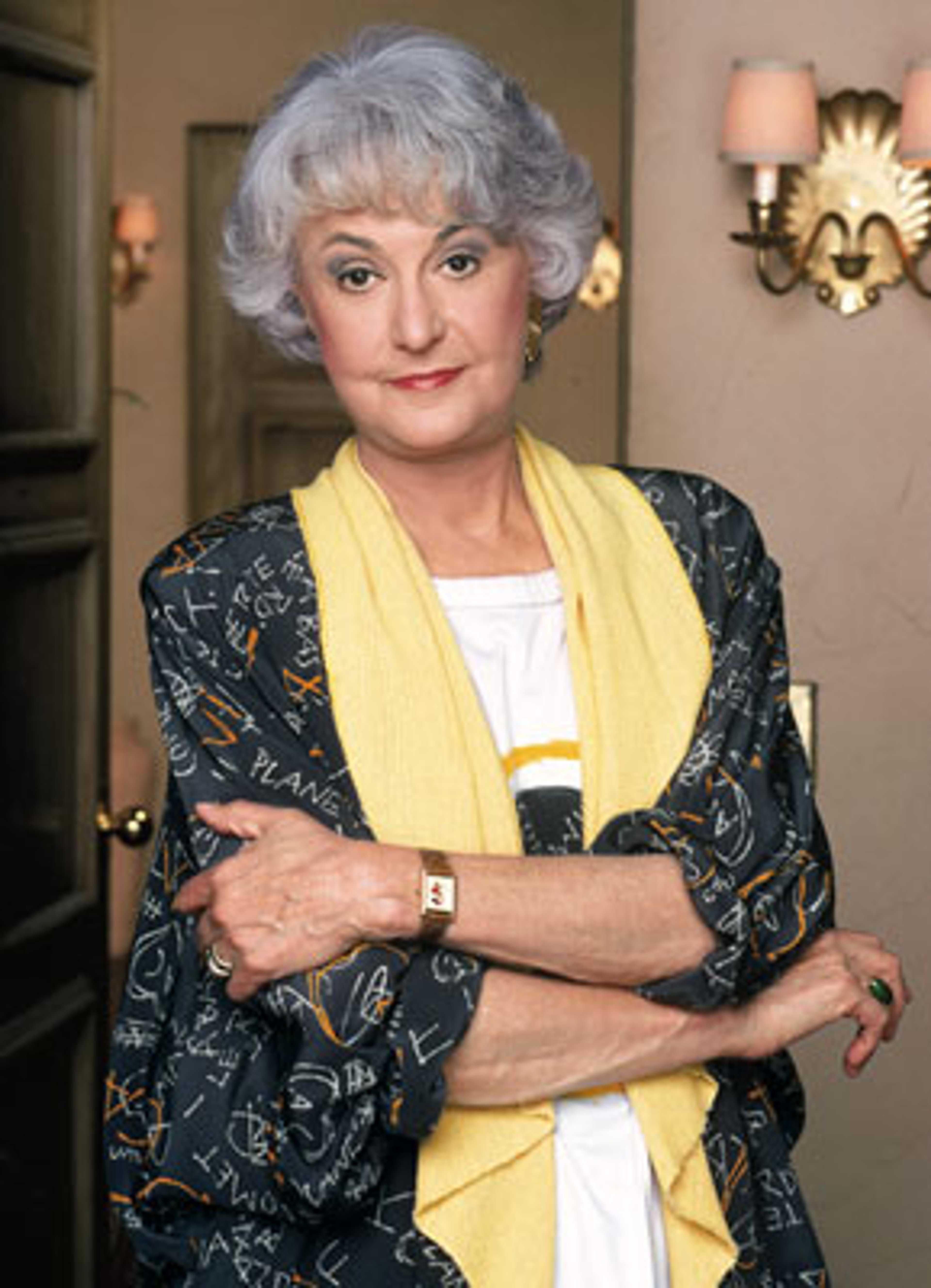 An older woman with gray curly hair wearing a black and white patterned blouse and a yellow scarf, looking directly at the camera.