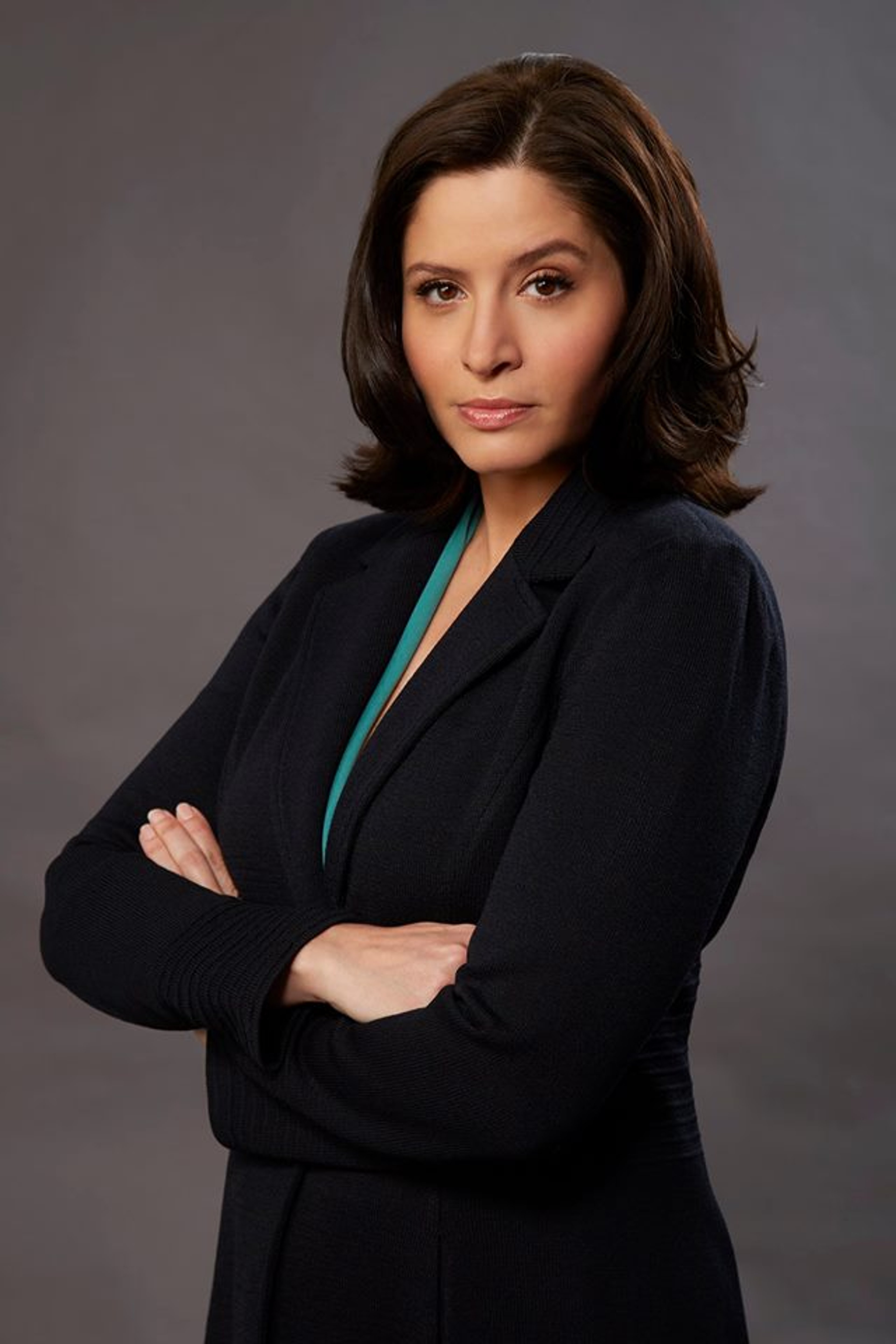 A woman with dark hair wearing a black coat, with a serious expression on her face and her arms crossed.