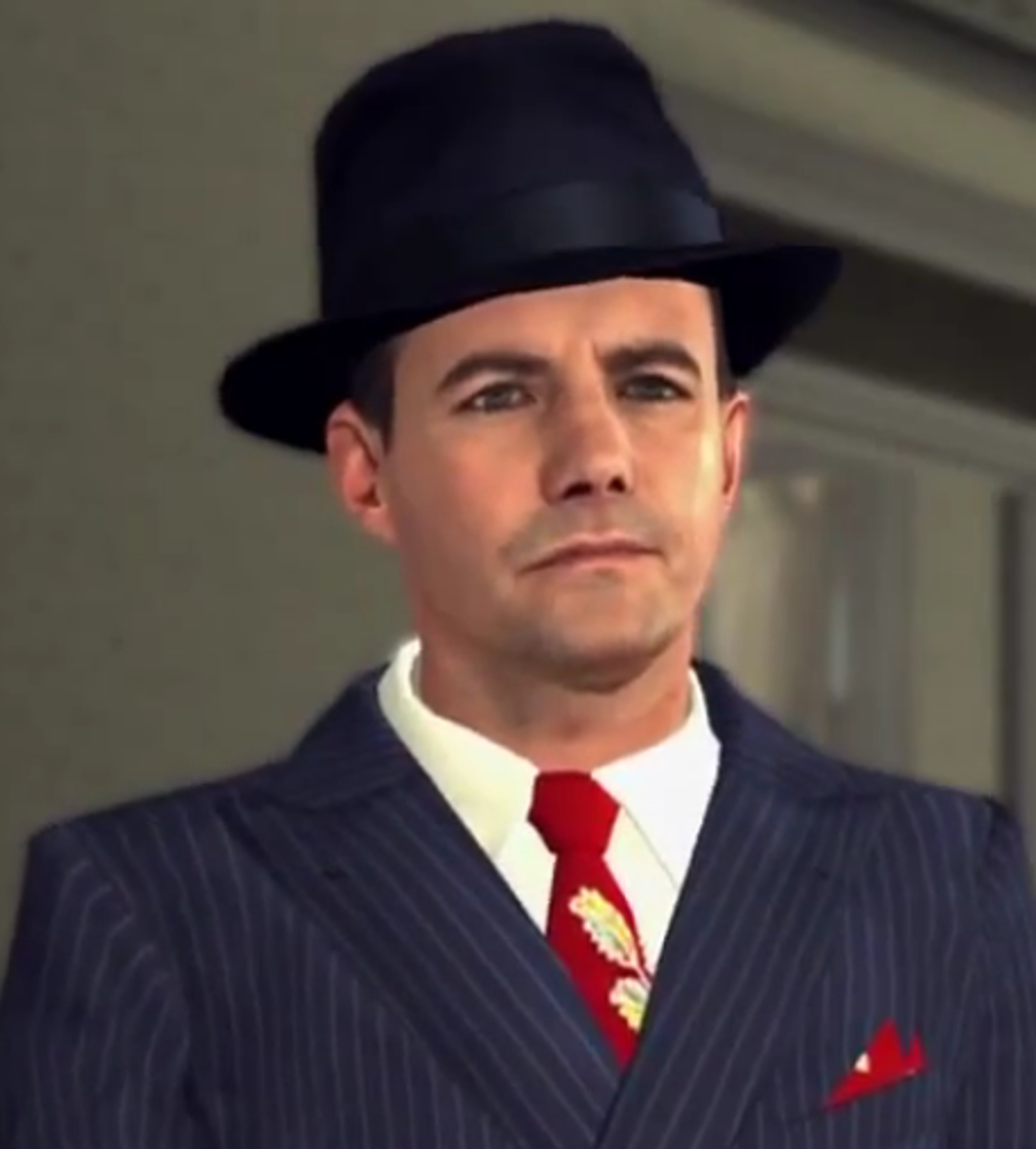 A middle-aged man in a suit and hat with a serious expression