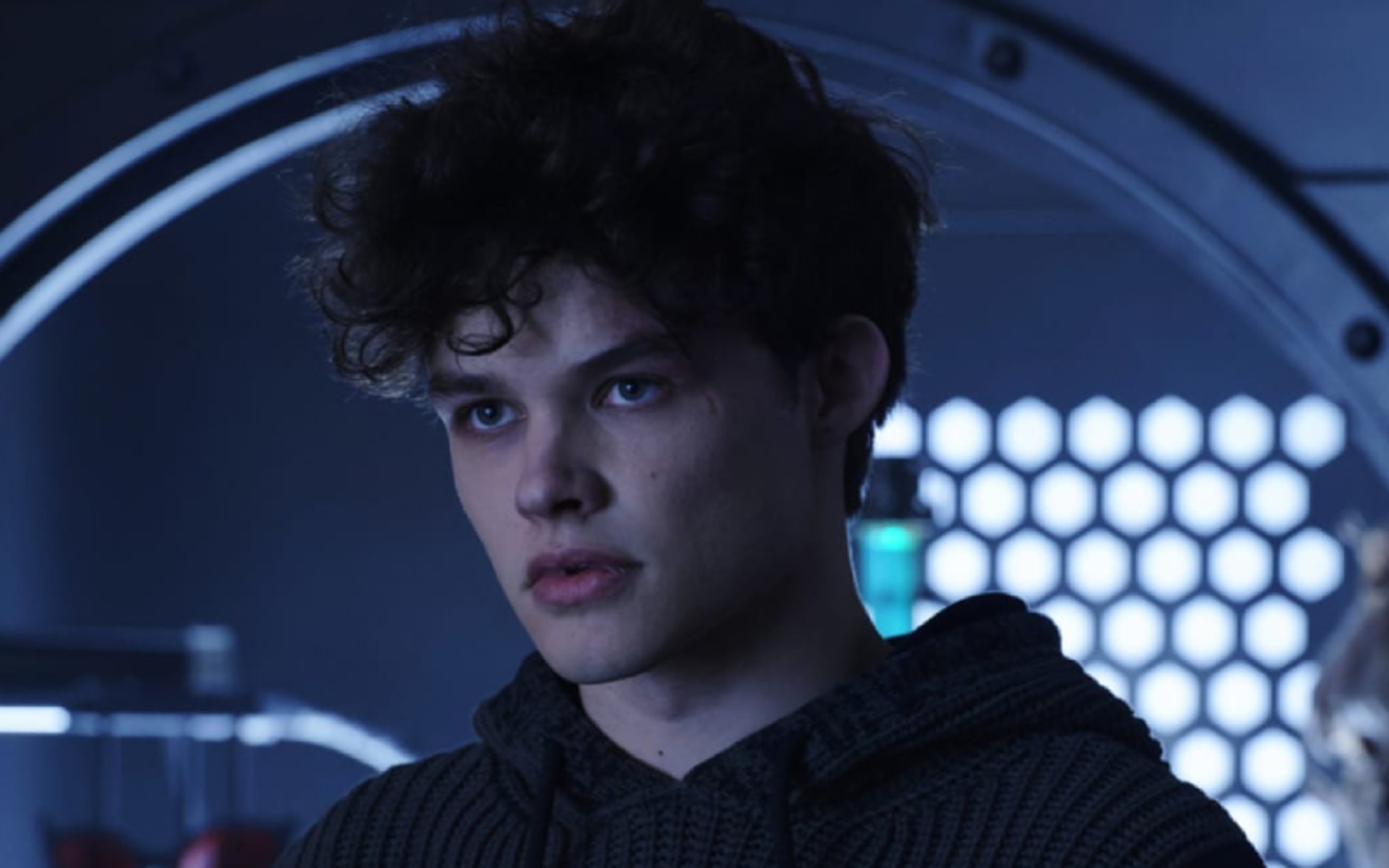 A young man with dark hair wearing a black jacket or shirt against a blue-tinted, futuristic background.