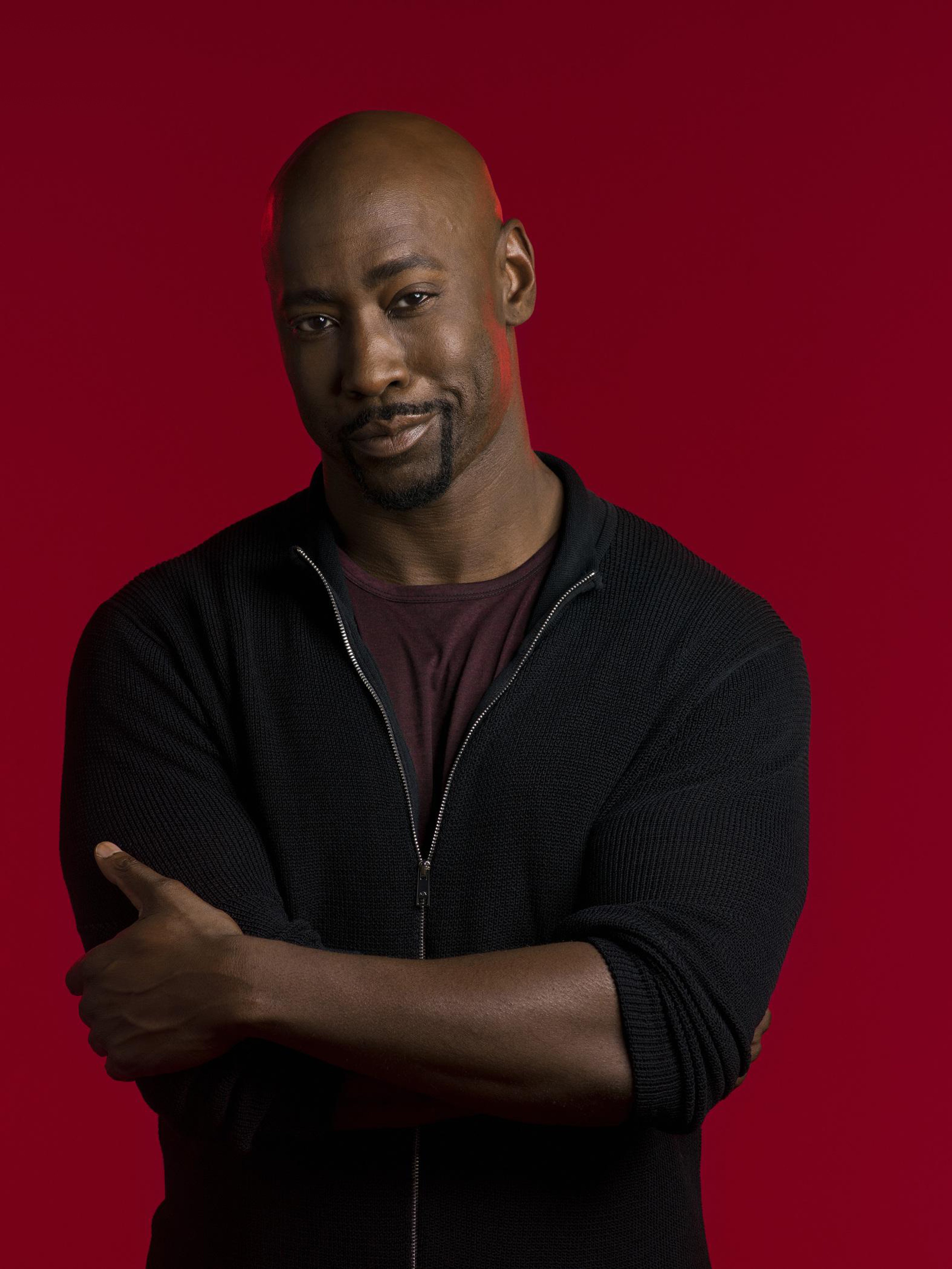 A serious-looking Black man with a muscular build wearing a black jacket