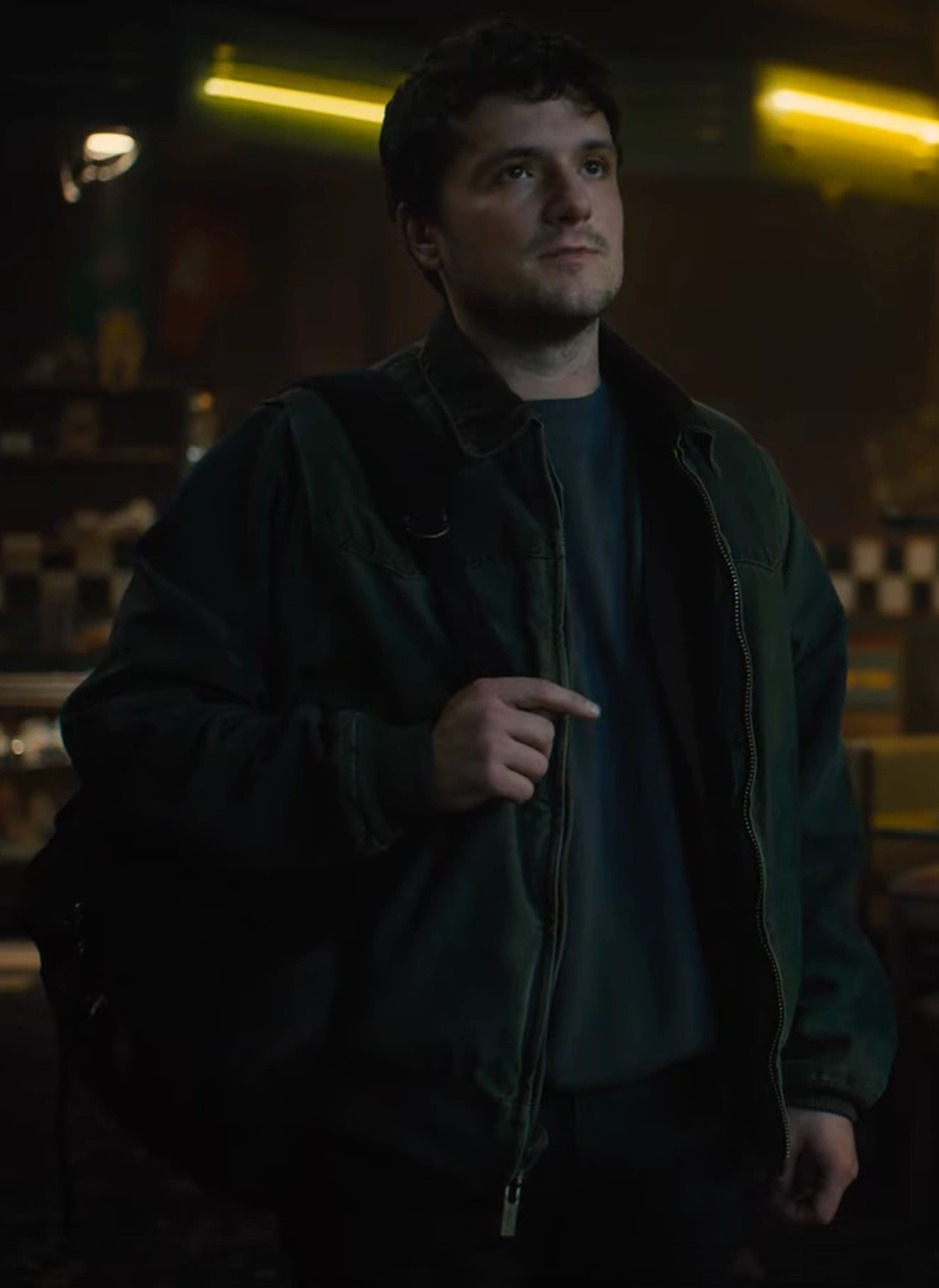 A young man with dark hair wearing a black jacket in a dimly lit environment.