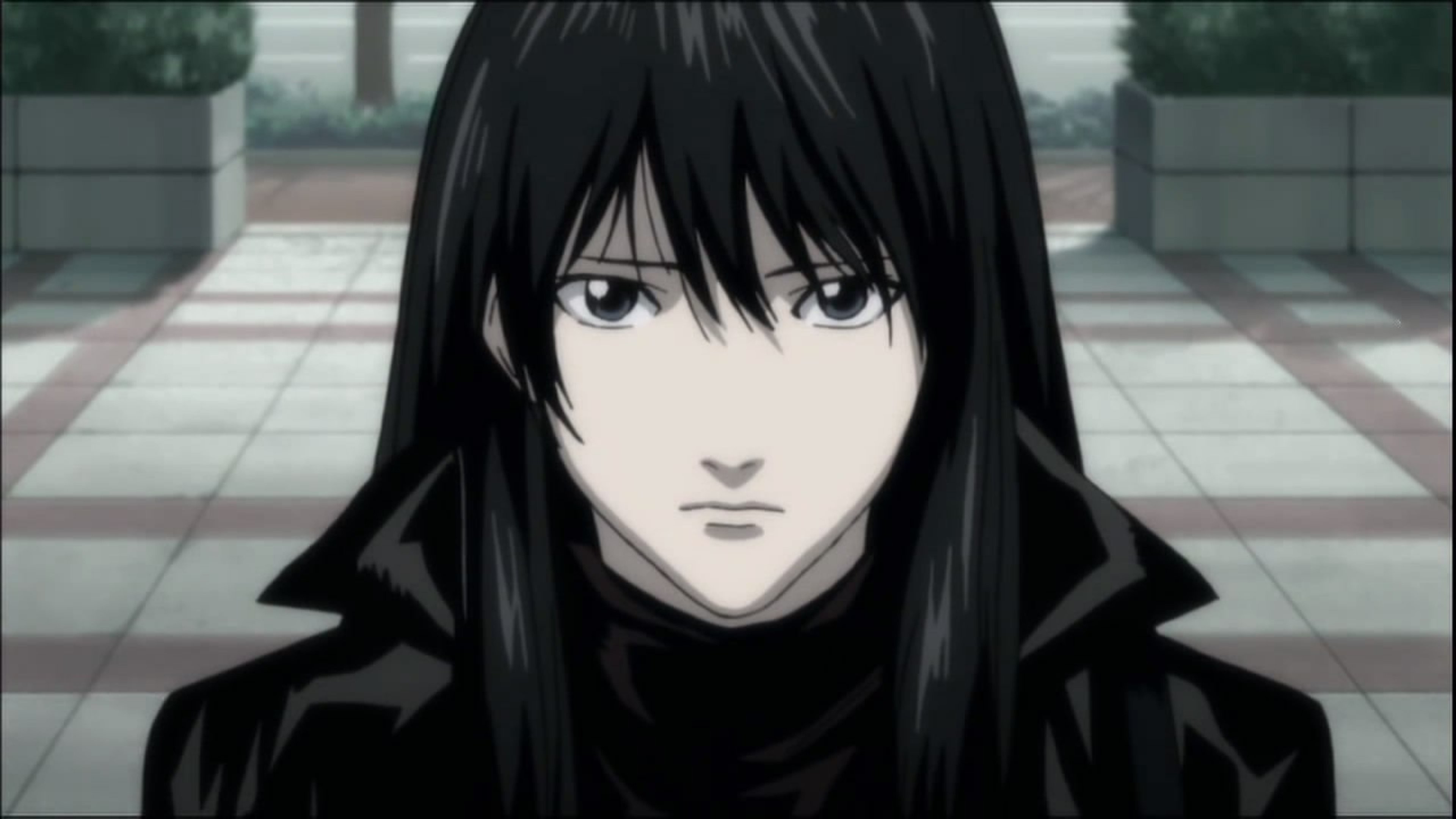 A young woman with dark hair wearing a black hooded jacket or cloak, with a serious expression on her face.