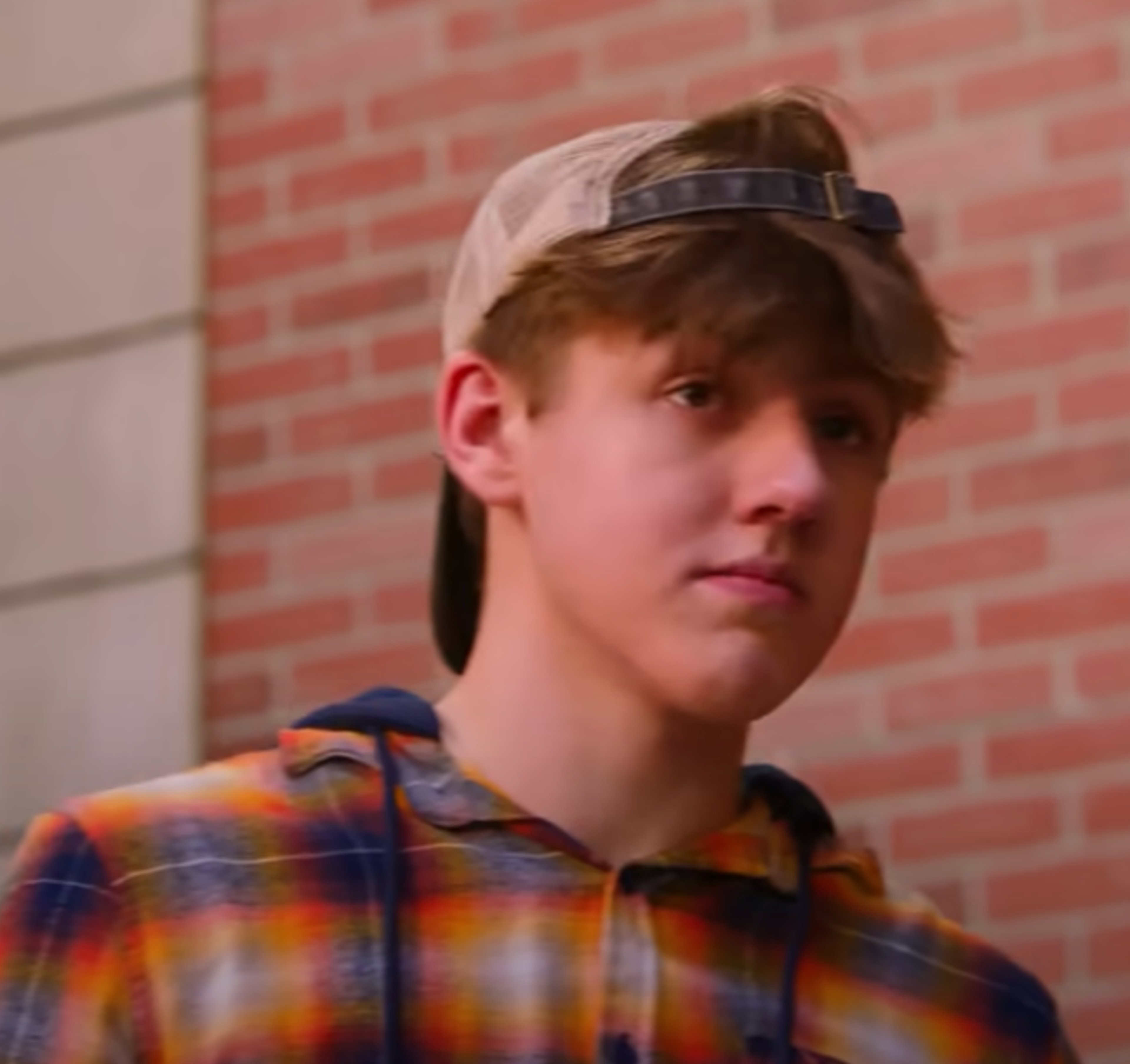 A teenage boy with short brown hair wearing a plaid shirt standing in front of a brick wall.