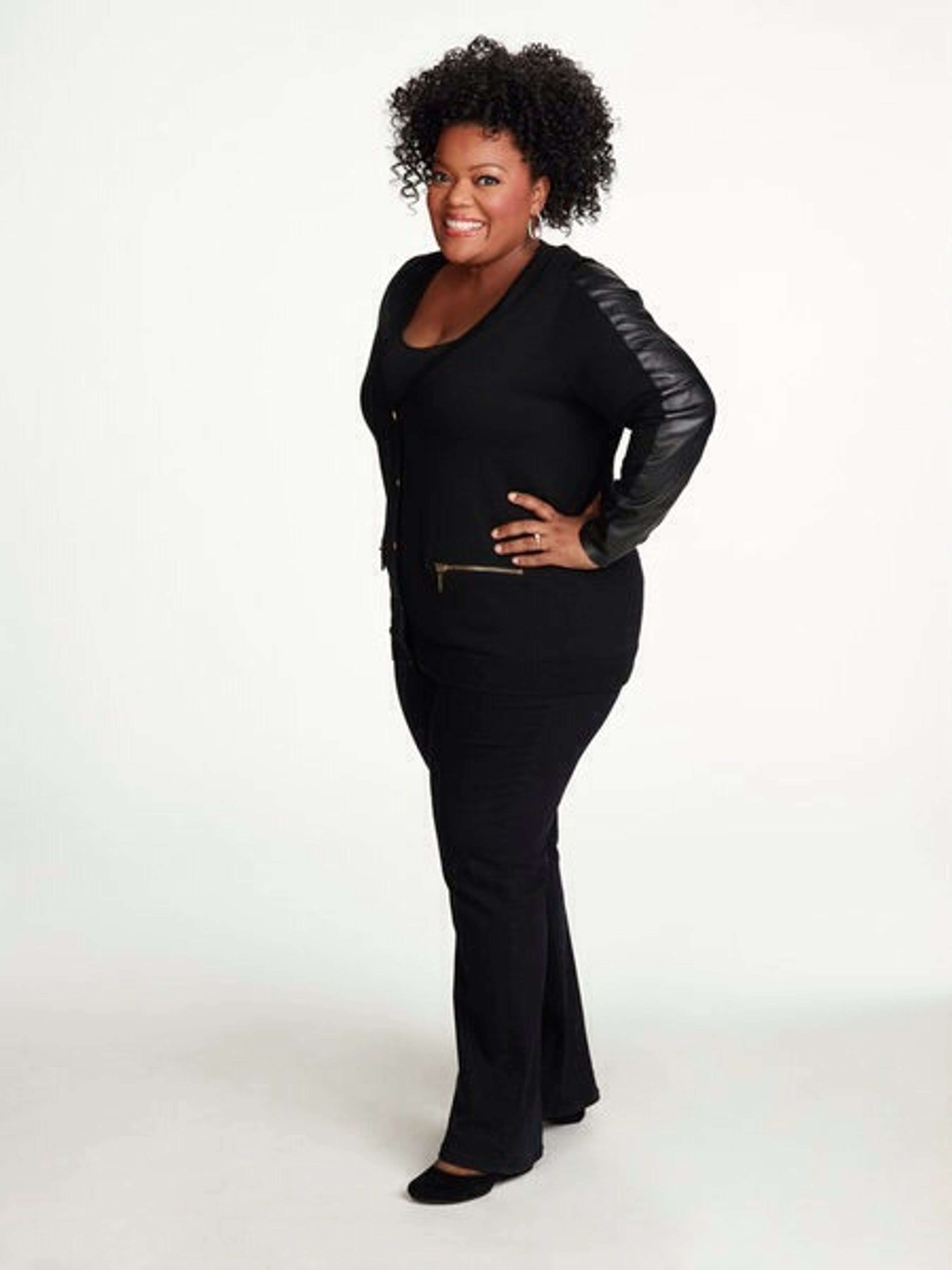 A middle-aged Black woman with curly hair wearing a black outfit, standing in a studio or office setting.