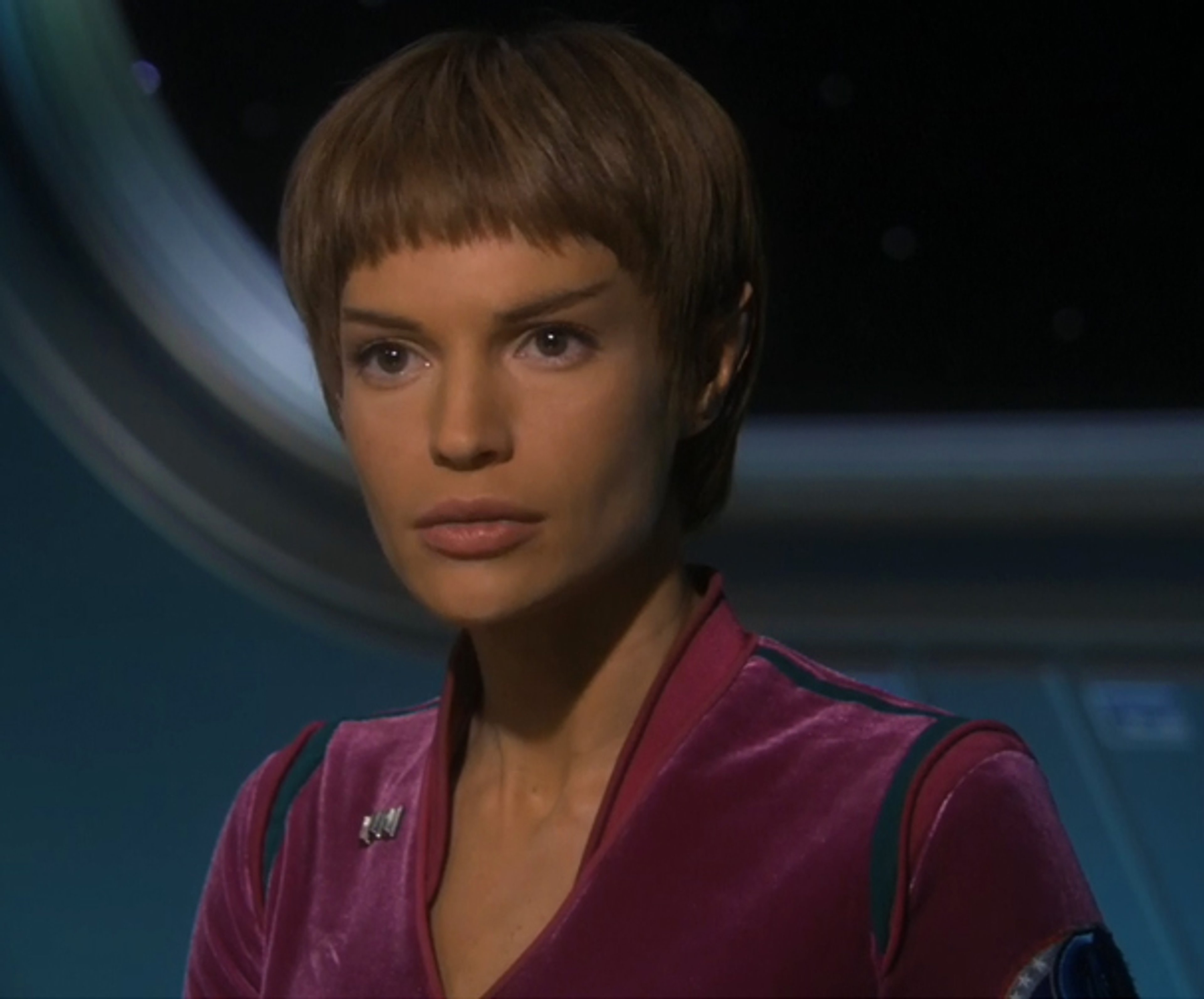 A woman with short brown hair in a bowl cut wearing a reddish-pink uniform, looking serious and focused.