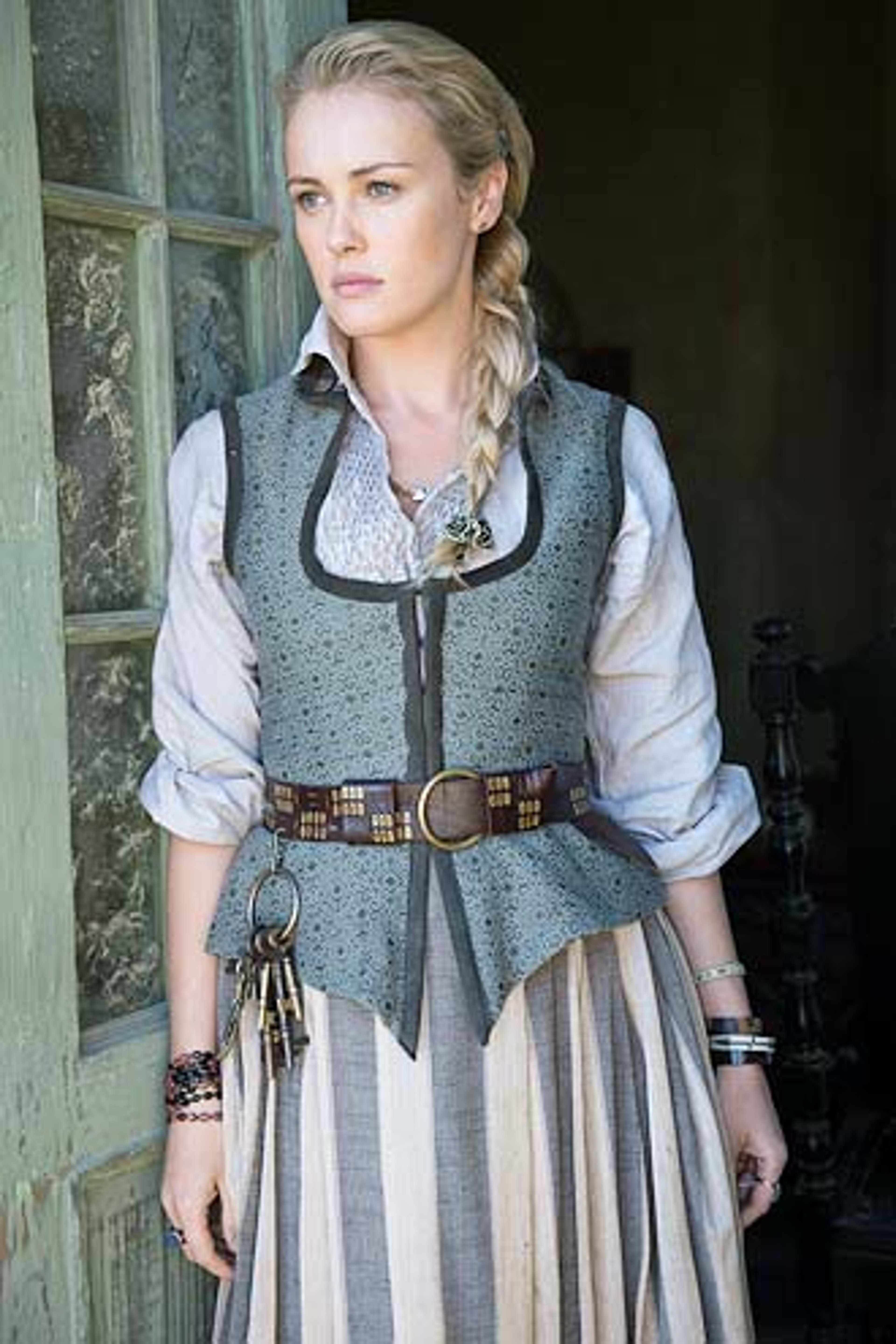 A young woman with braided blonde hair wearing a gray vest and striped skirt, standing in front of a wooden building.