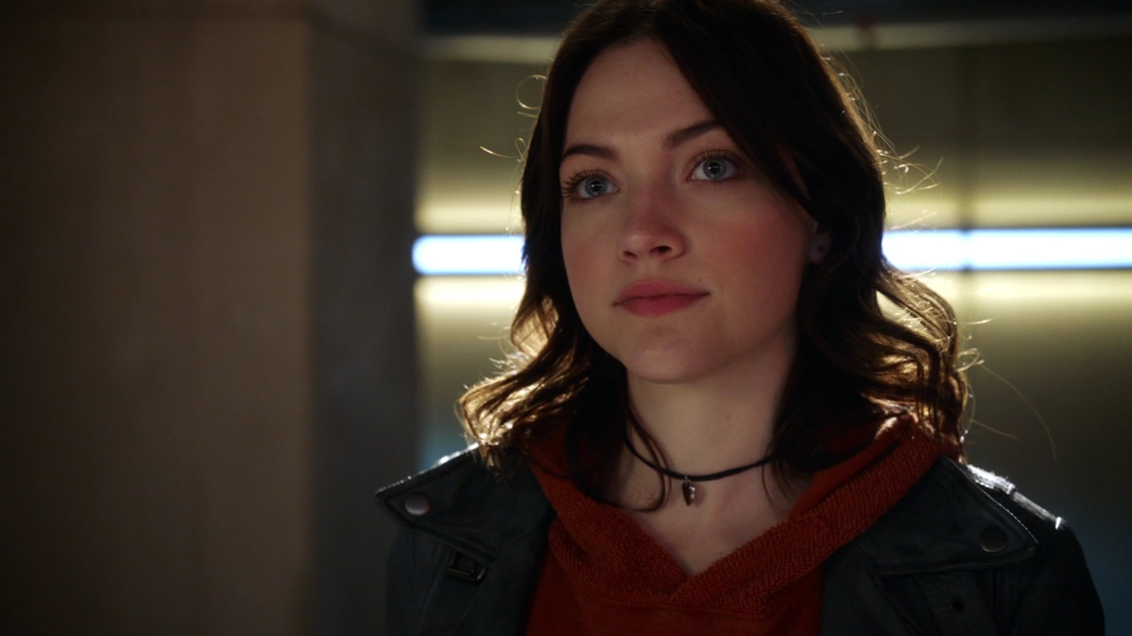 A young woman with dark hair wearing a red jacket in a dimly lit indoor setting.