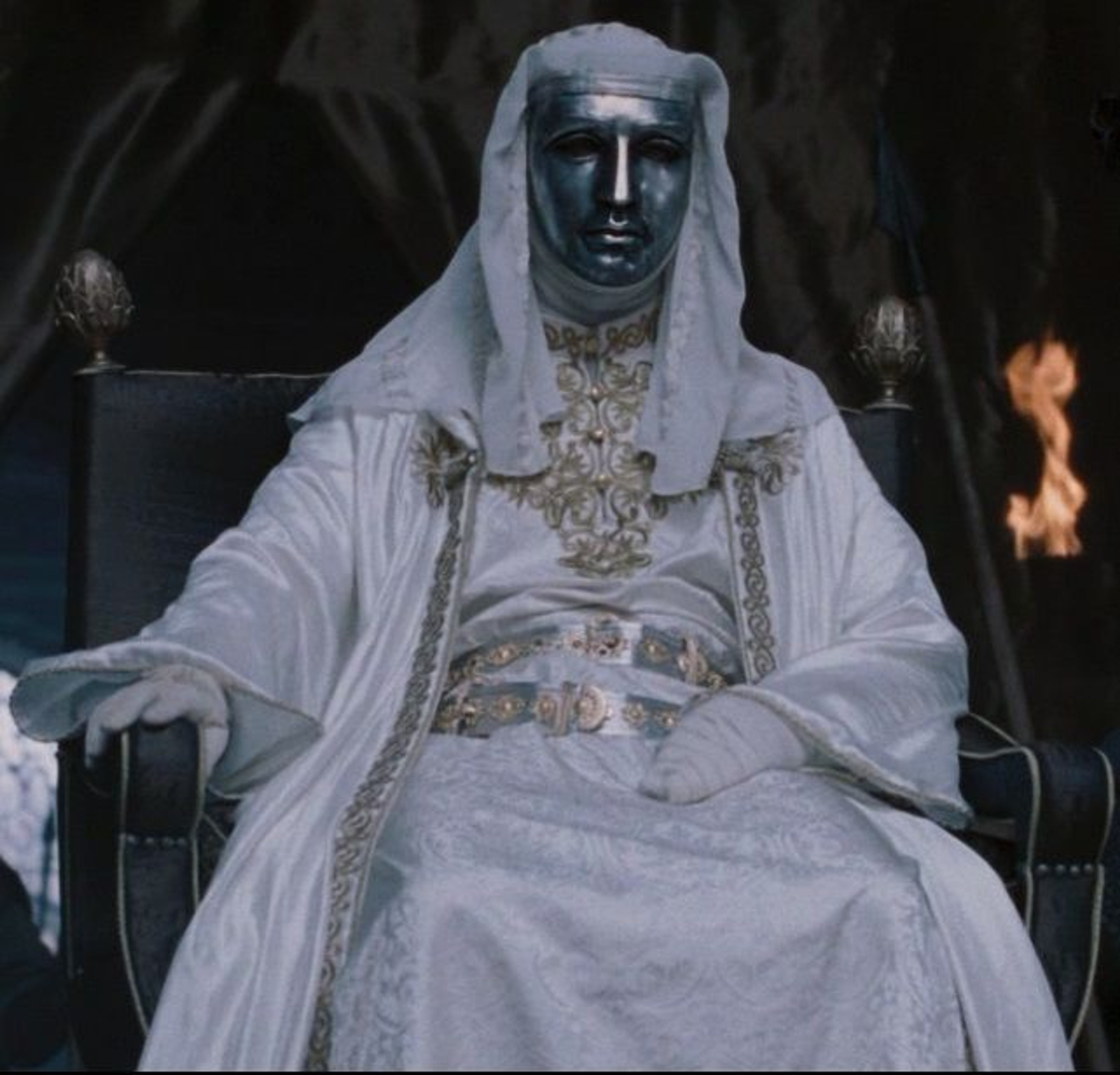 A man wearing a white robe and headdress, with a mask covering the lower half of his face, sitting on a throne or chair.