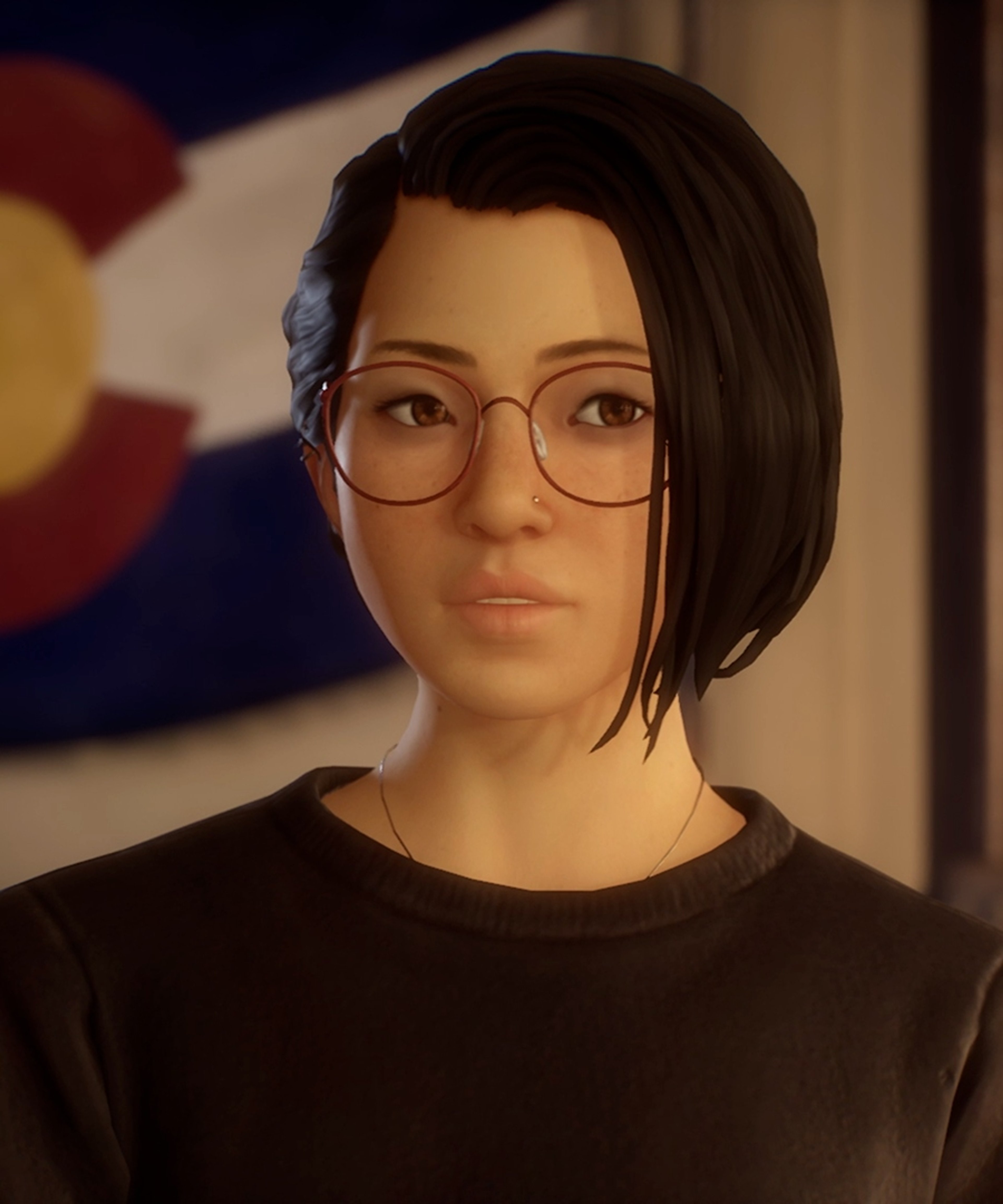 A young woman with short dark hair and glasses, wearing a black shirt and looking serious.