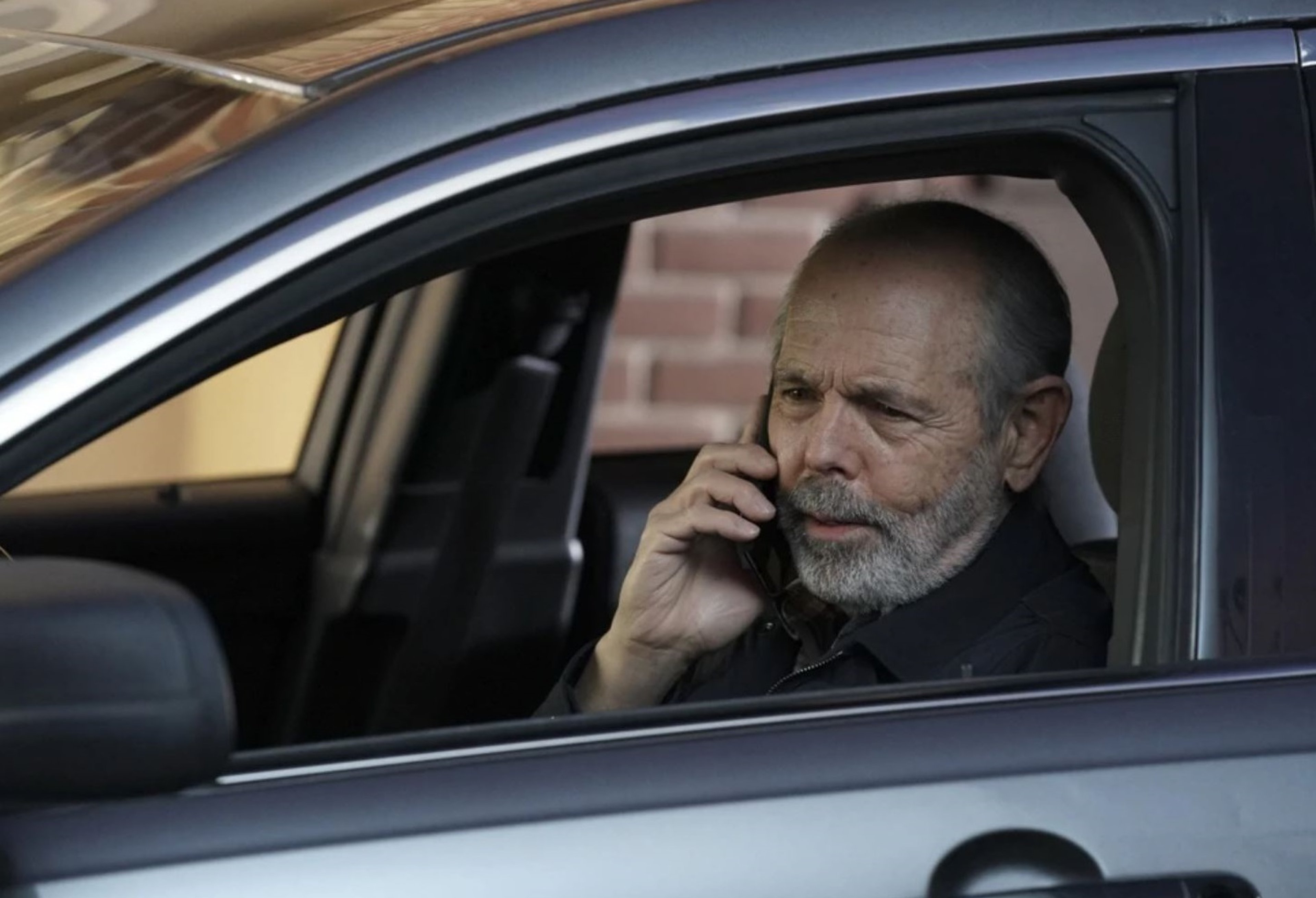 A middle-aged man with gray hair and a beard sitting in a car and talking on a cell phone.