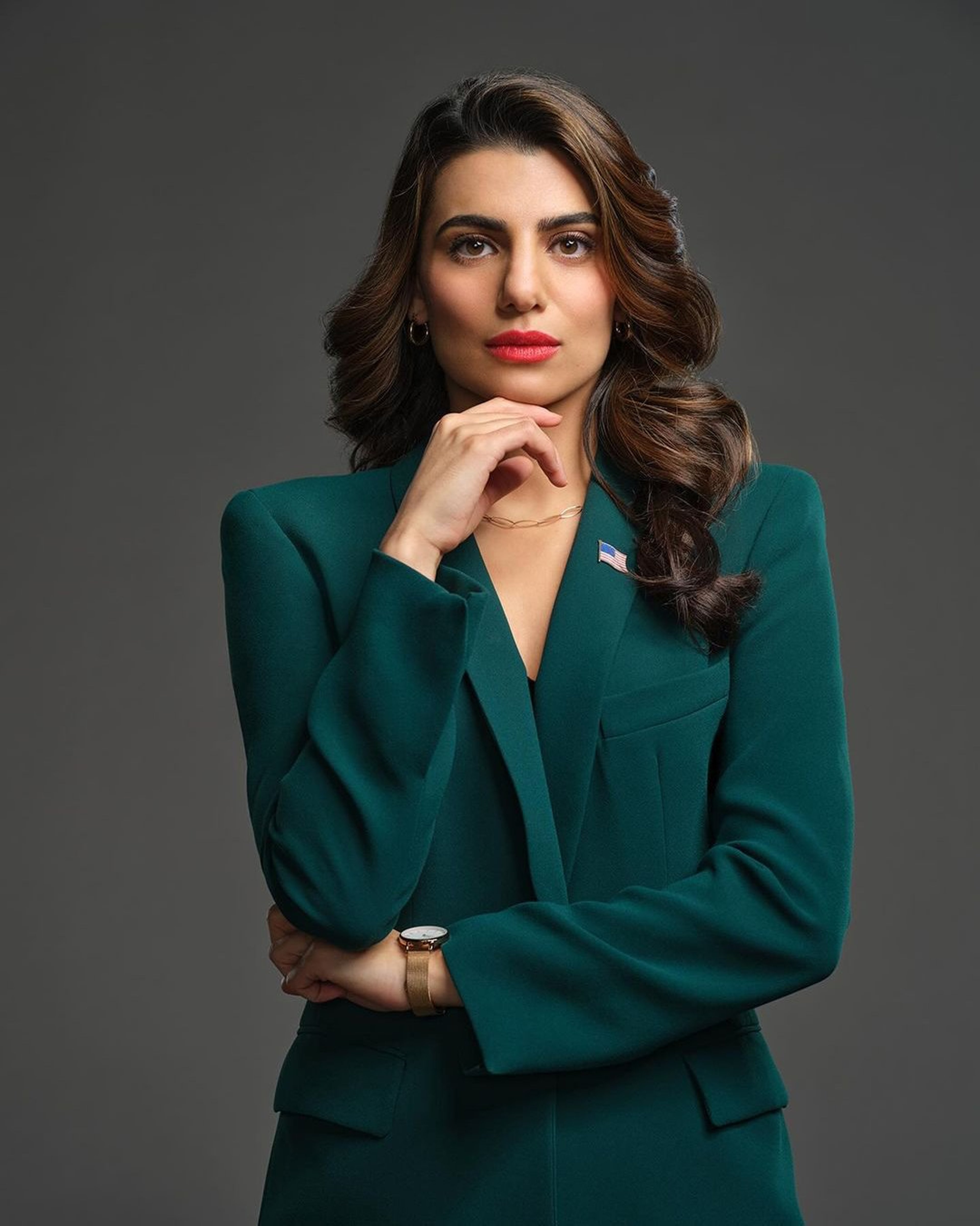 A woman with long, dark curly hair wearing a green blazer and looking directly at the camera.