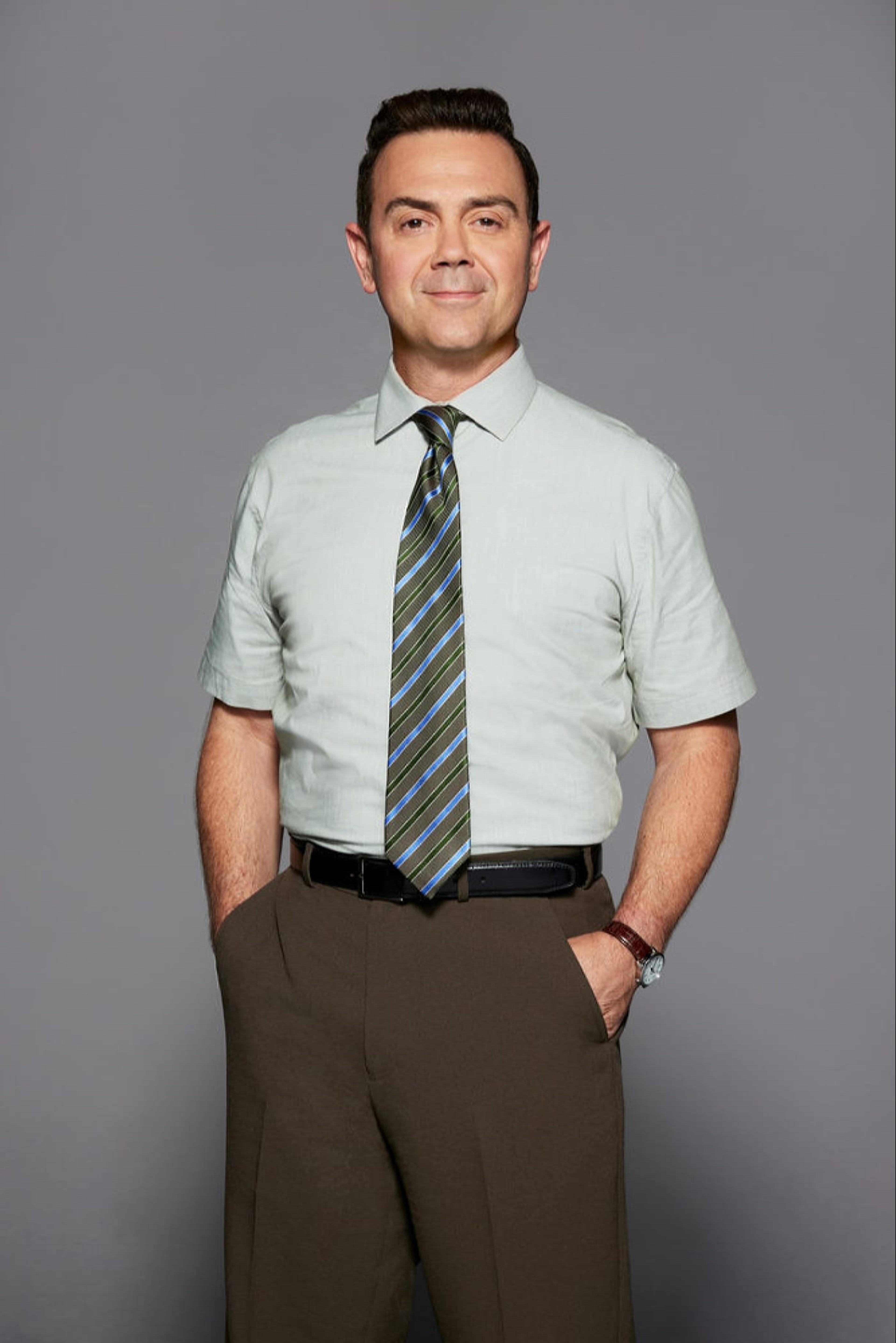 A middle-aged man in a light blue shirt and tie, standing with his hands in his pockets.