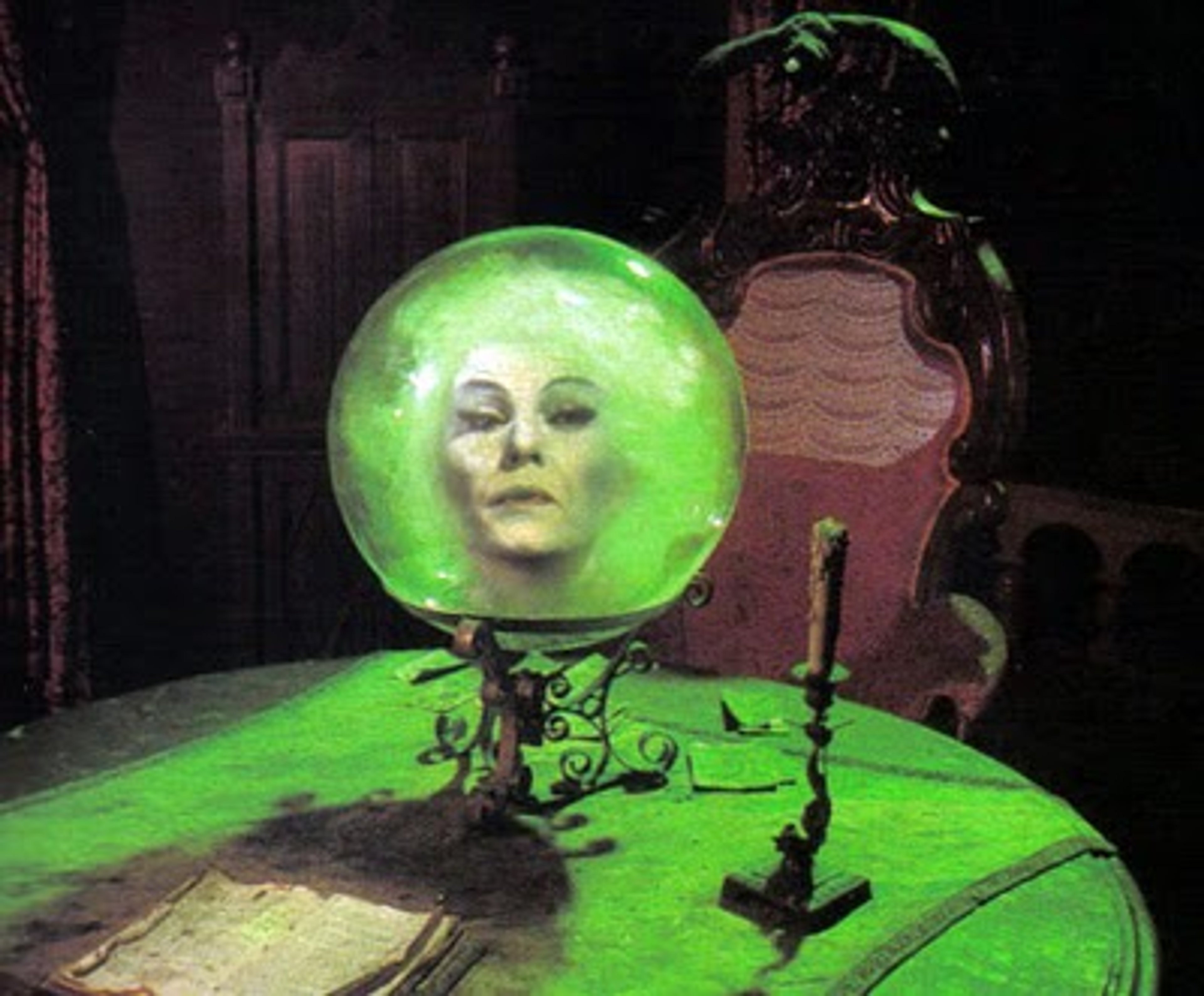 A close-up view of a glowing green crystal ball containing the face of a mysterious, intense-looking woman