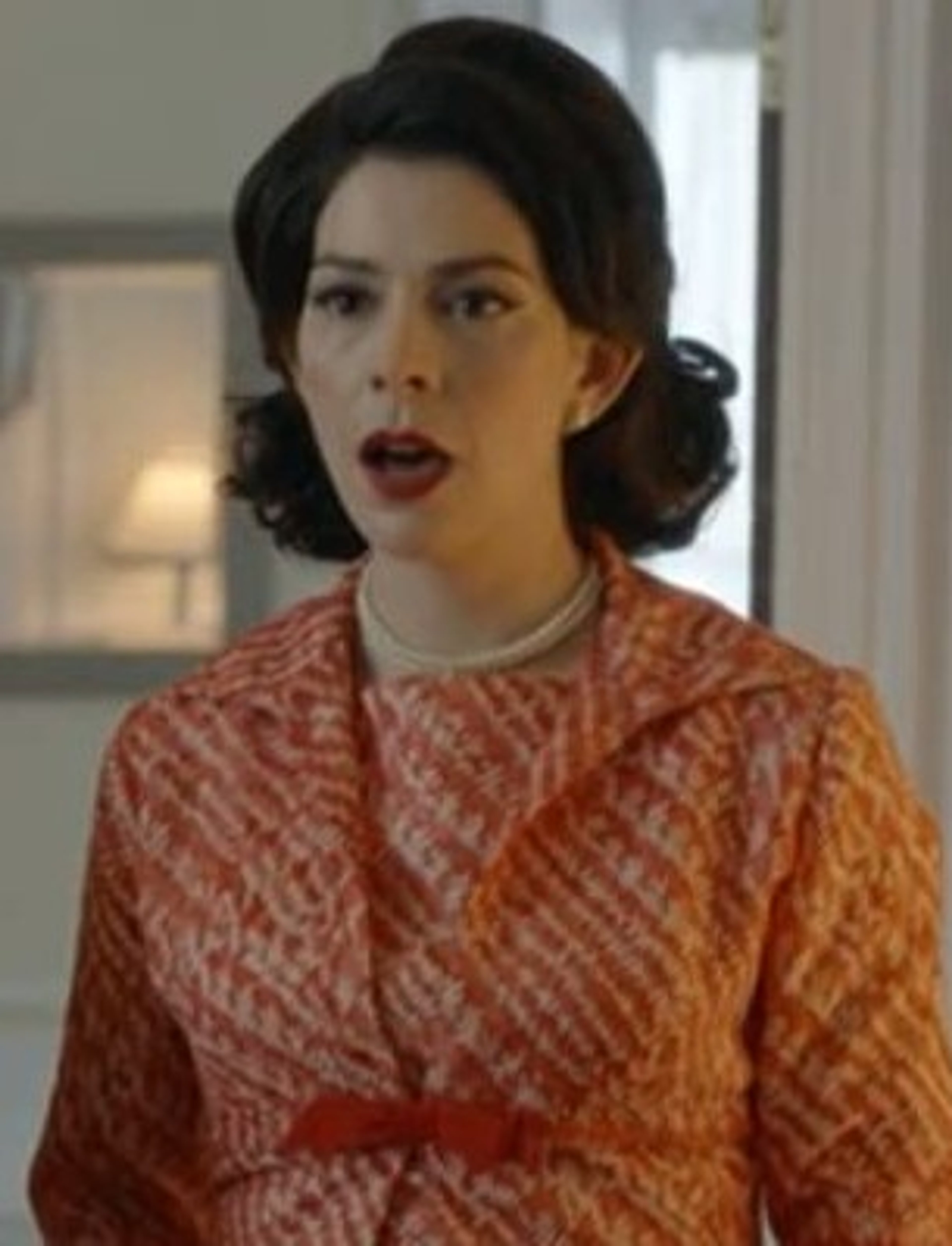 A woman with dark hair in an updo, wearing a red and white patterned dress, with a serious expression on her face.