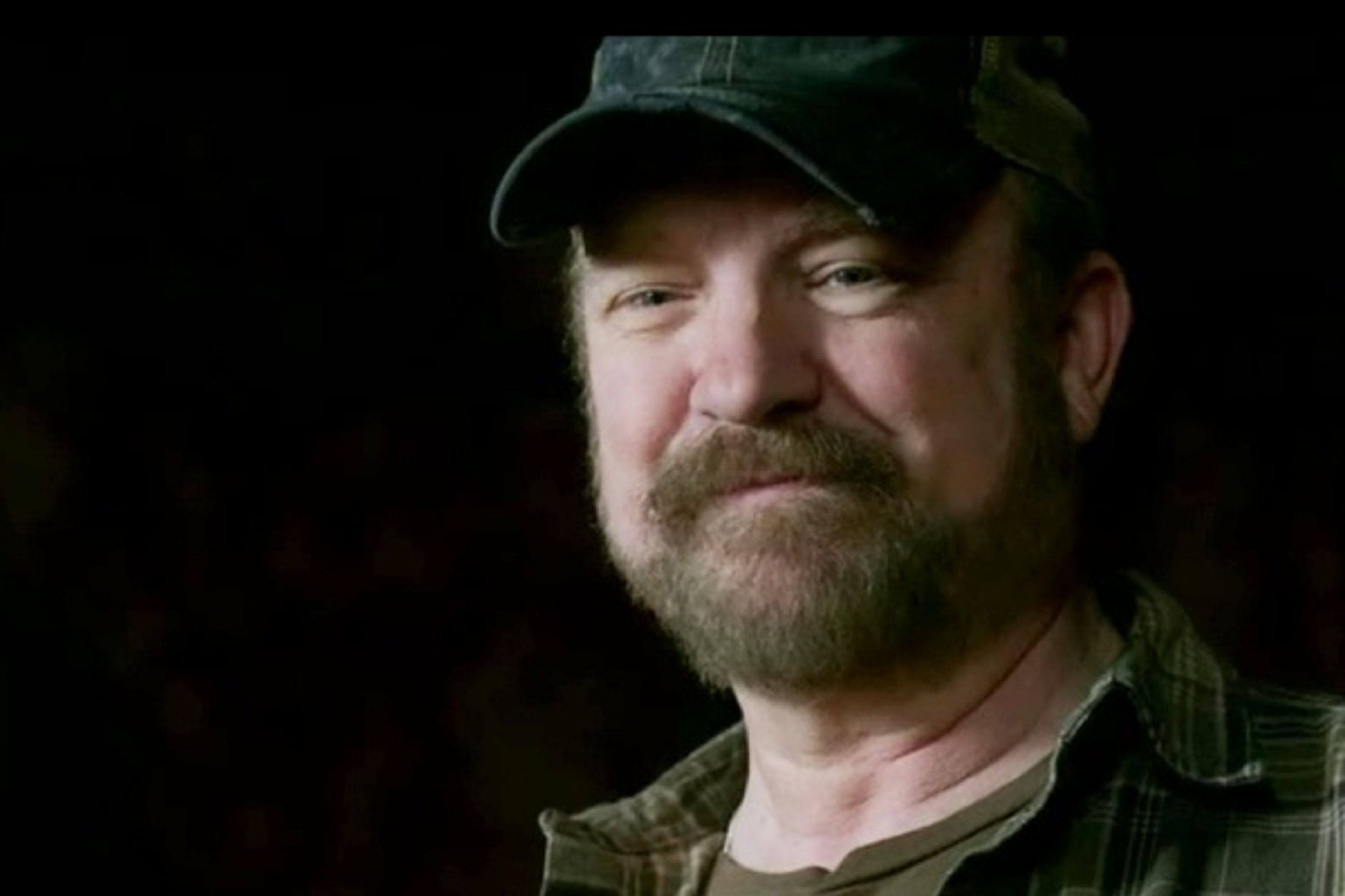 A middle-aged man with a beard wearing a baseball cap and plaid shirt