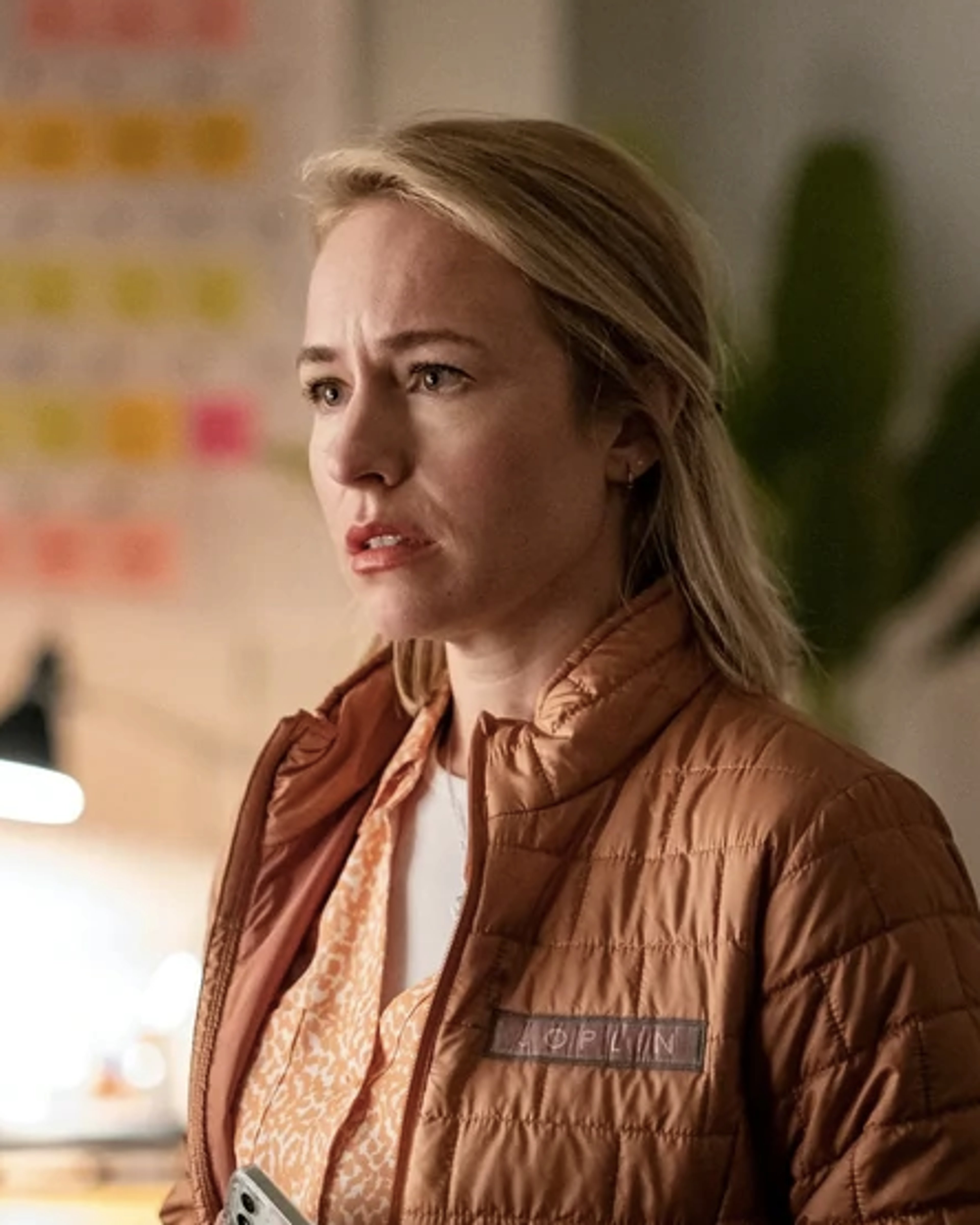 A middle-aged woman with blonde hair wearing a brown jacket in an office setting.