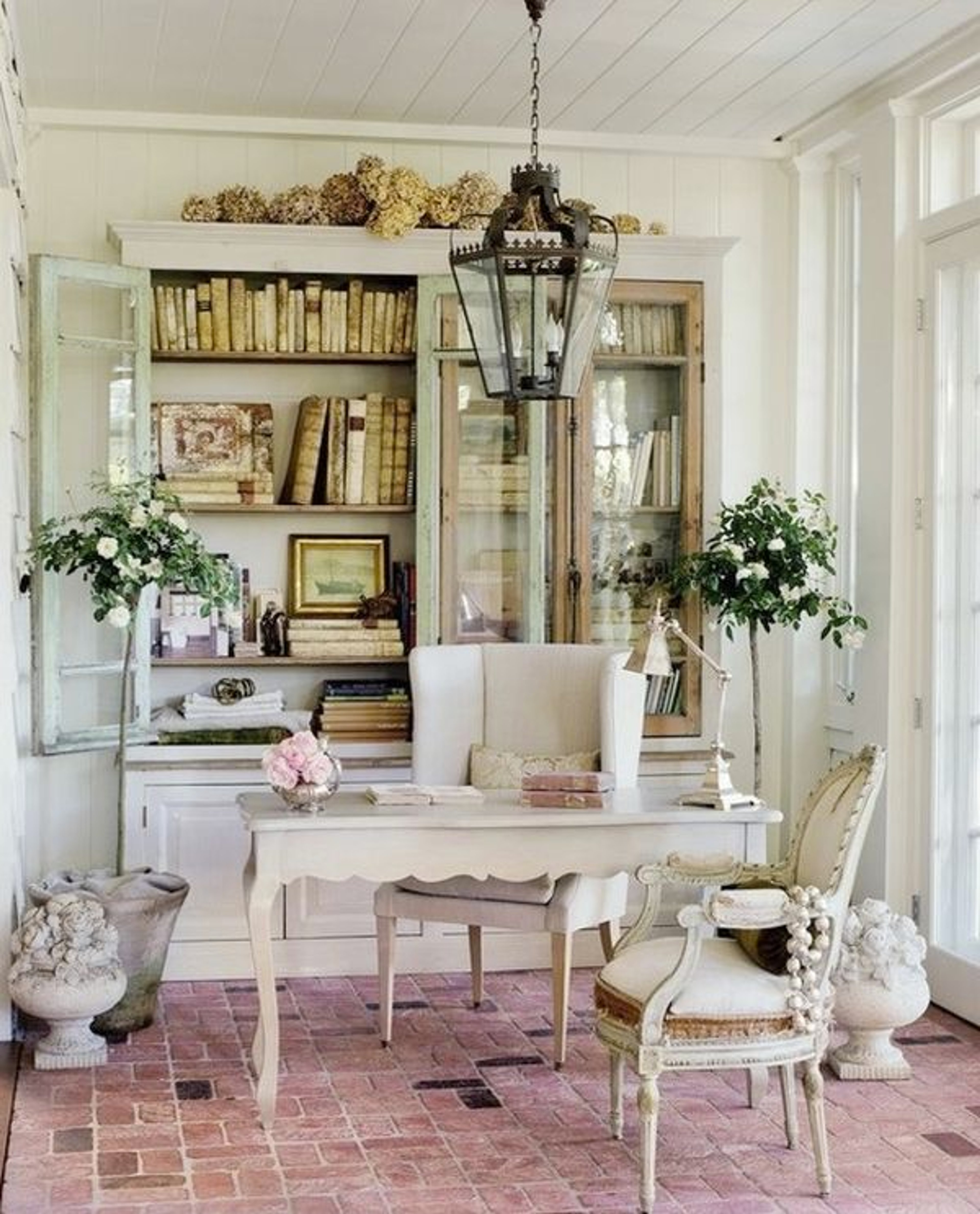 A cozy, feminine interior design space with a white wooden table, vintage chandelier, and bookshelves filled with books and decor.