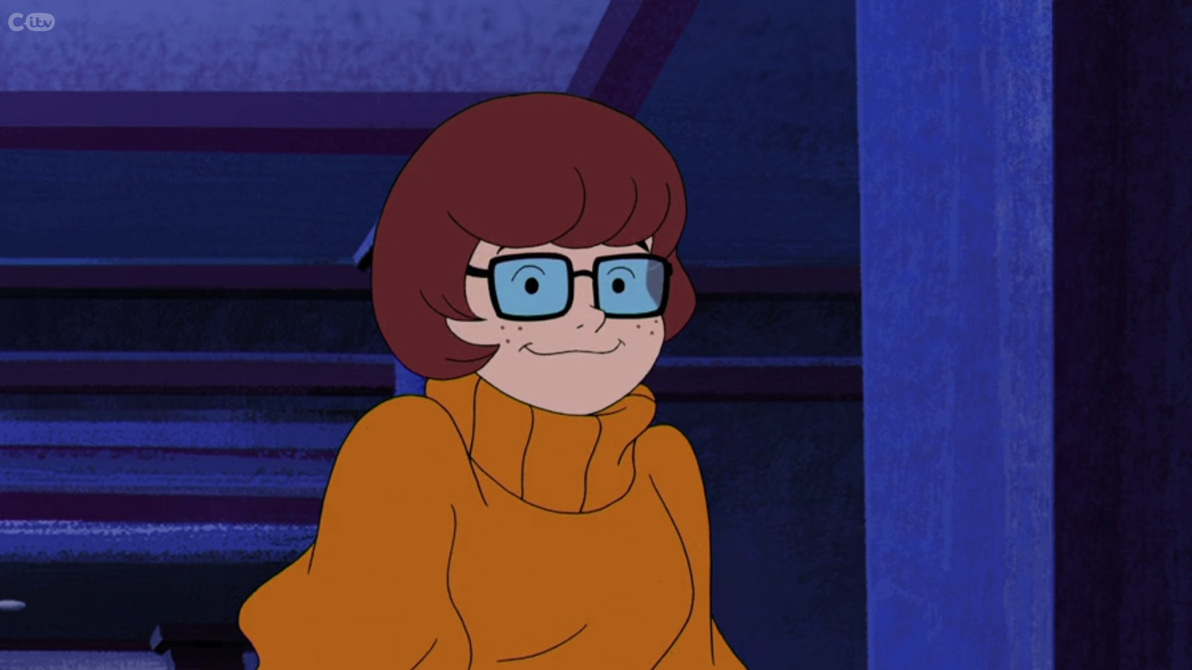A cartoon character with orange hair, glasses, and a brown sweater sitting in a chair.