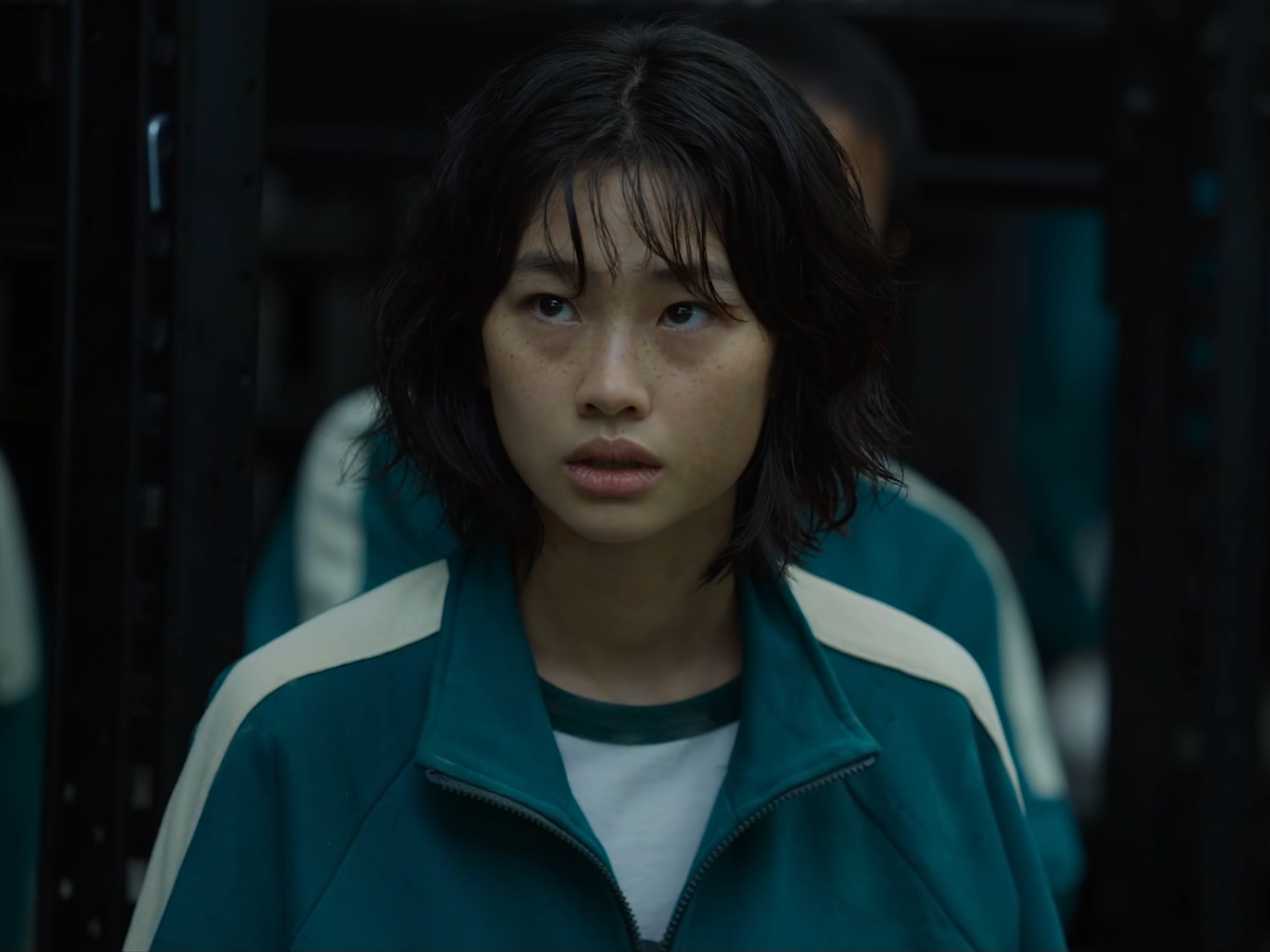 A young woman with short dark hair wearing a green jacket in a dimly lit, industrial setting.