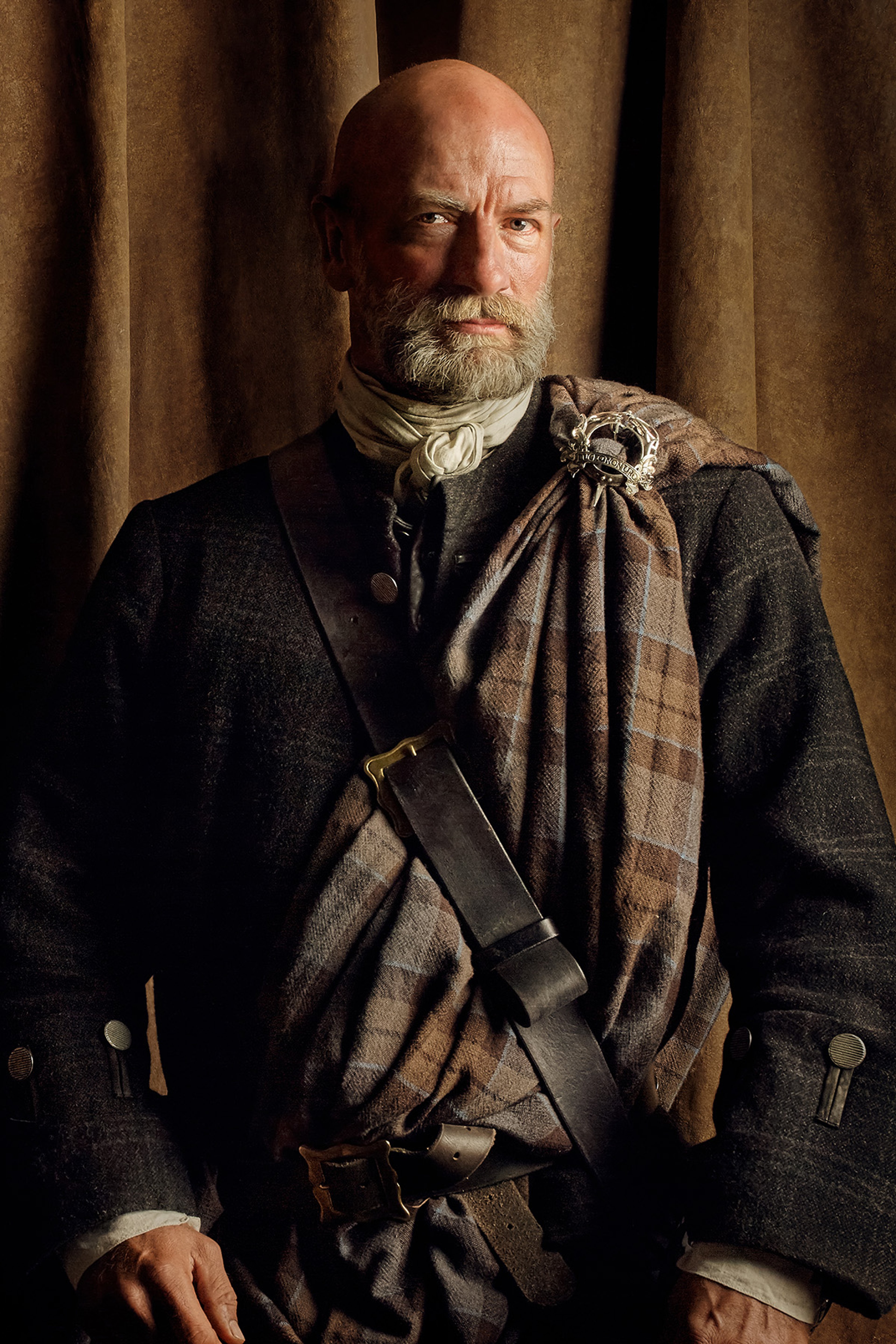 A rugged, older man with a beard wearing a dark coat and tartan scarf