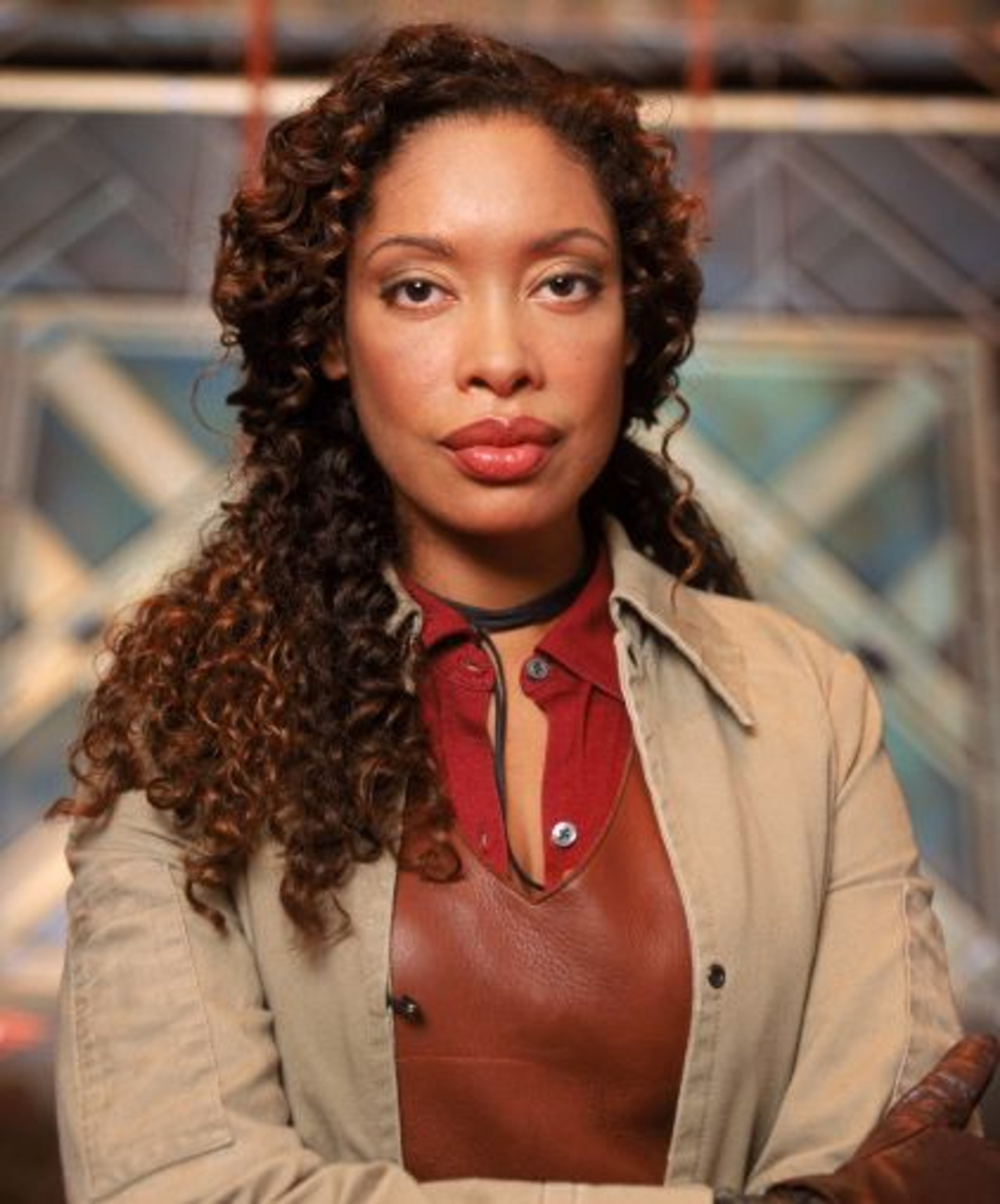 A woman with curly dark hair wearing a beige trenchcoat, looking serious and determined.