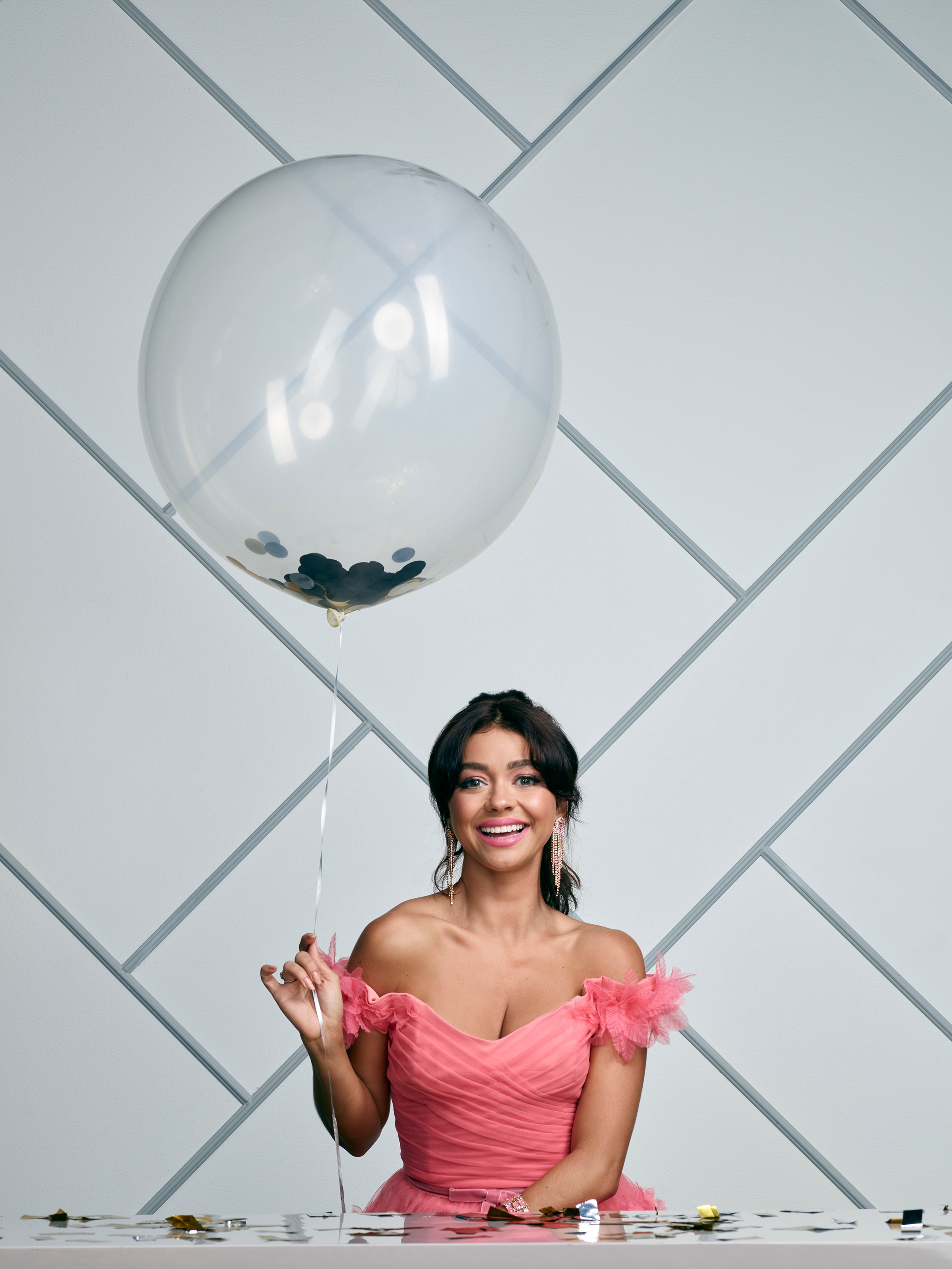 A young woman in a pink dress holding a transparent balloon in front of a white geometric patterned wall
