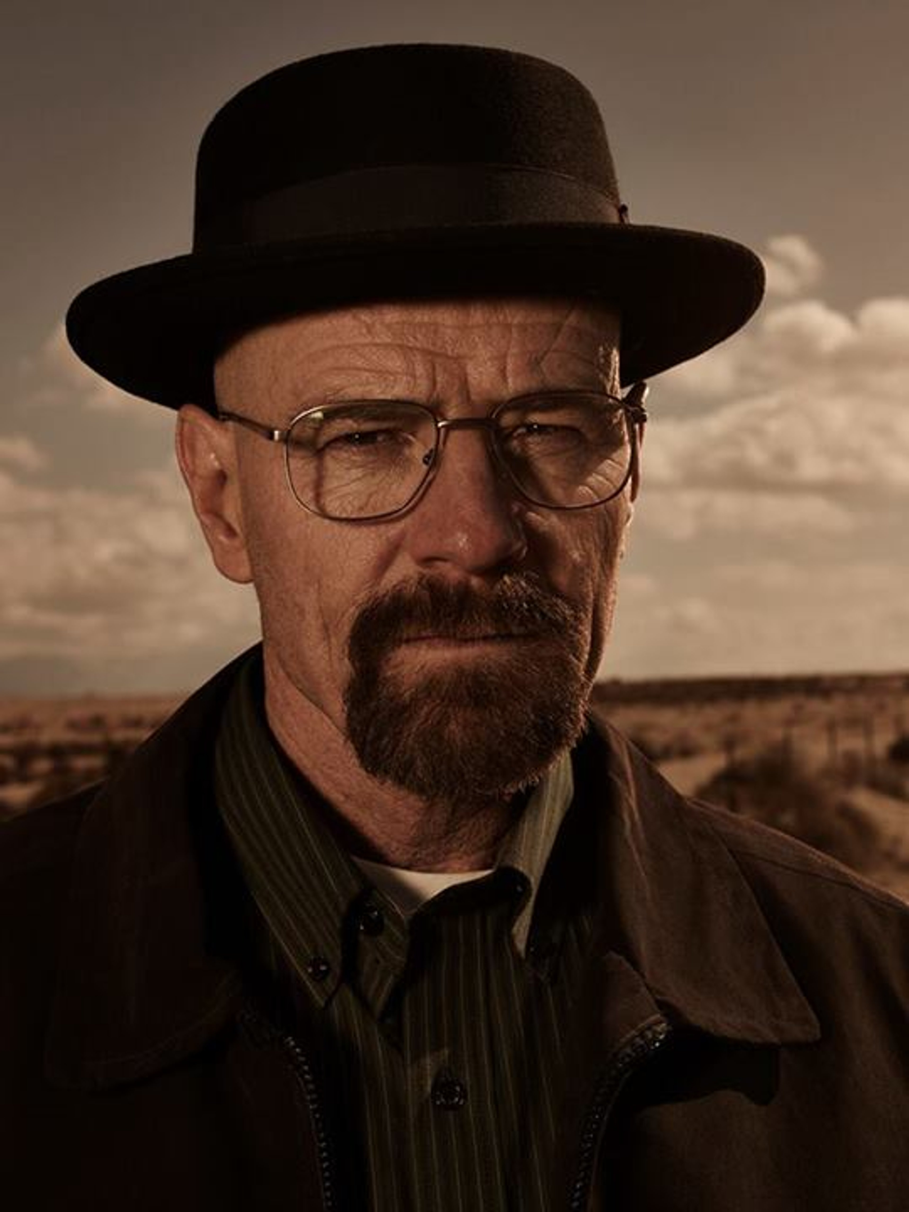 A middle-aged man with a beard wearing a black hat and glasses, standing in a desert or rural landscape