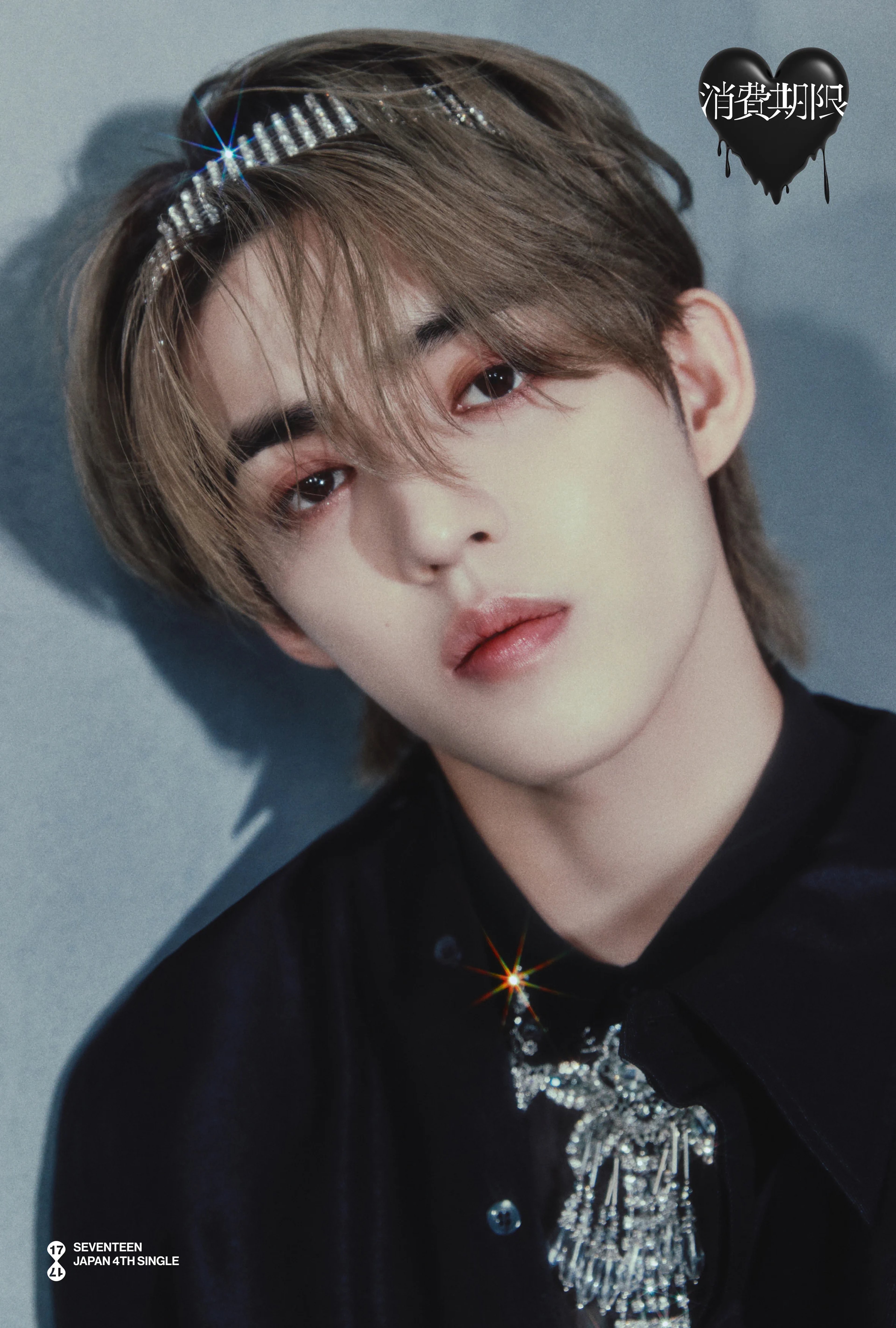 A young man with dark hair and a serious expression, wearing a black shirt and a silver hair accessory.