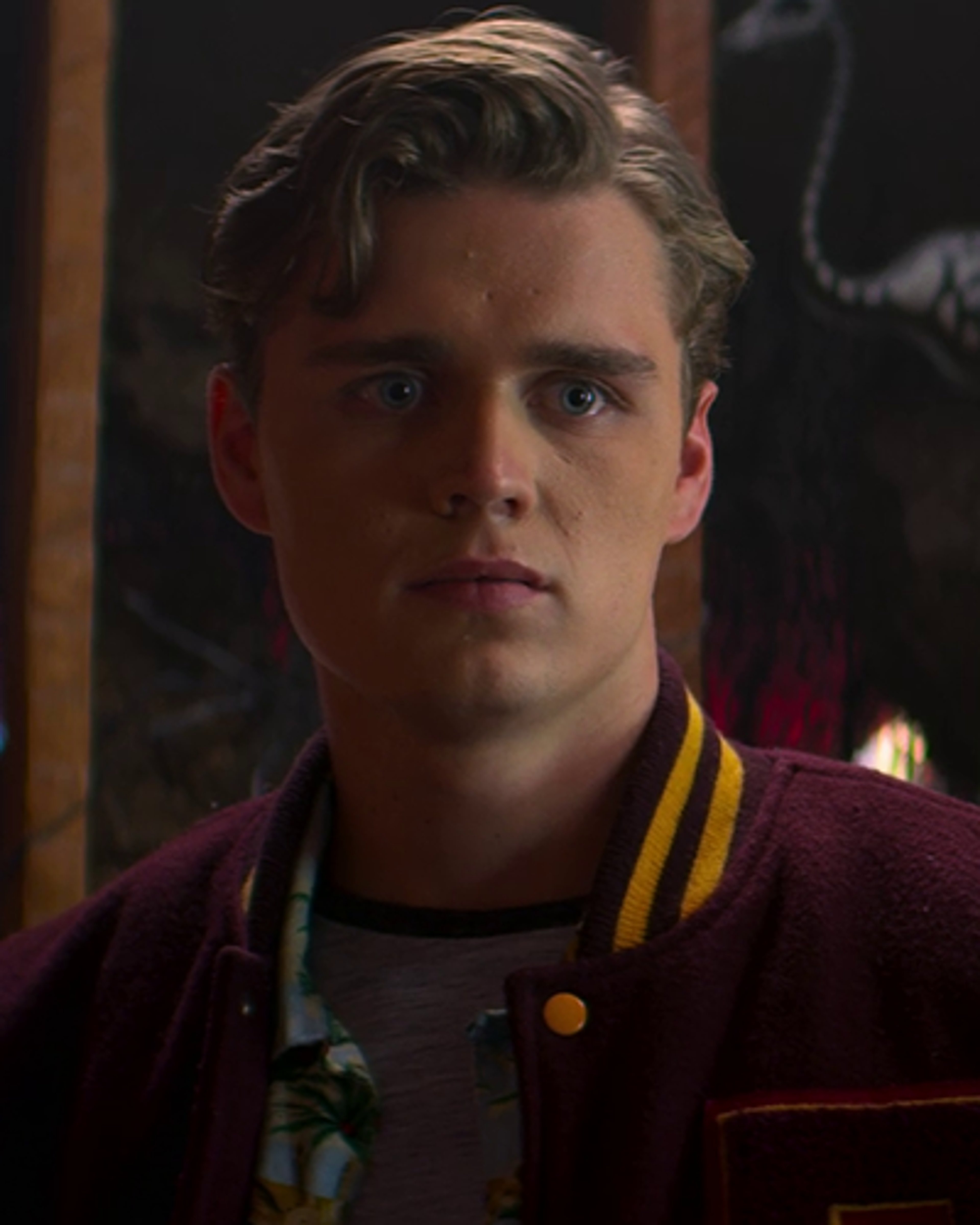 A young man with wavy blonde hair wearing a maroon jacket with yellow stripes