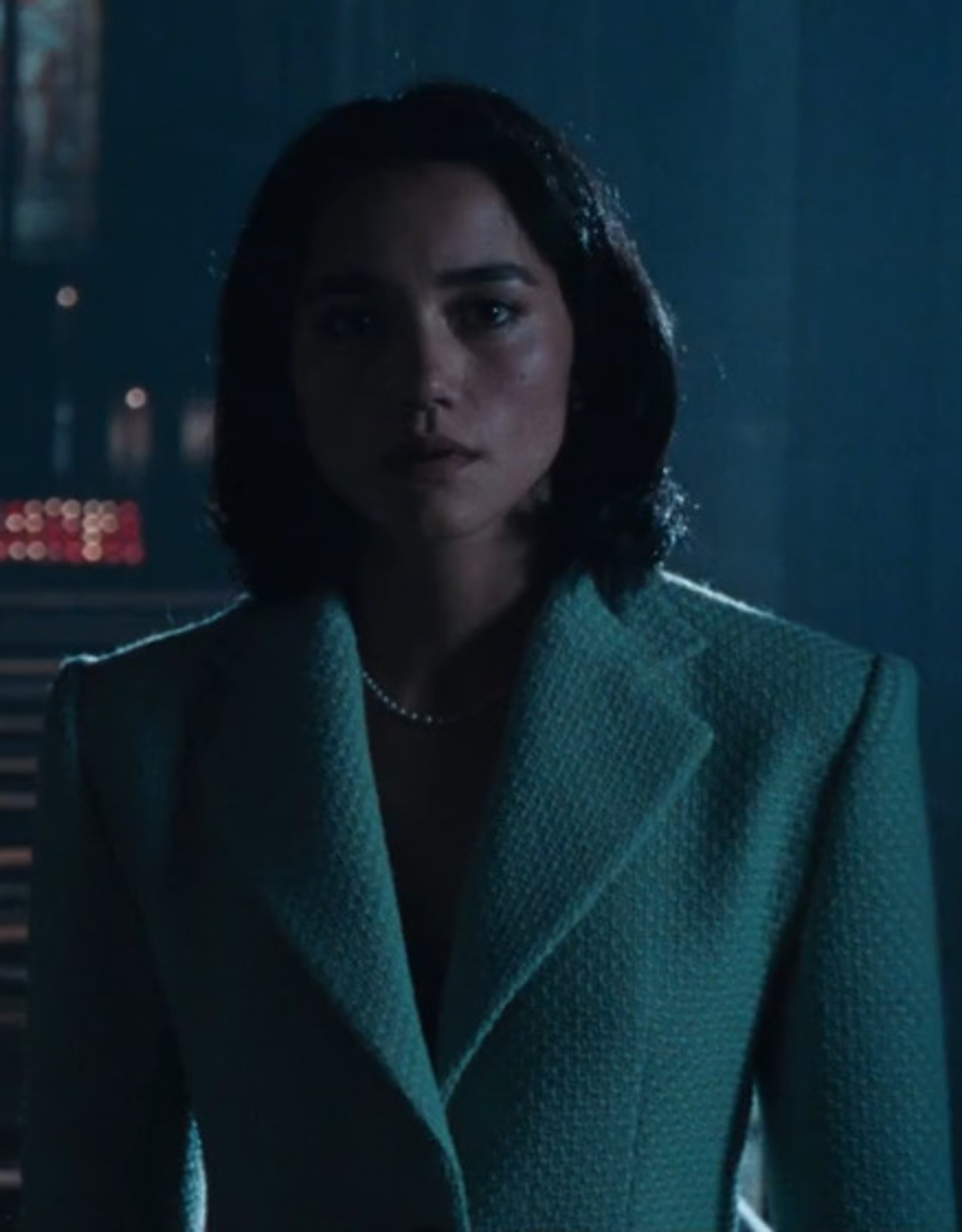 A woman with wavy brown hair and a green coat in a dimly lit setting