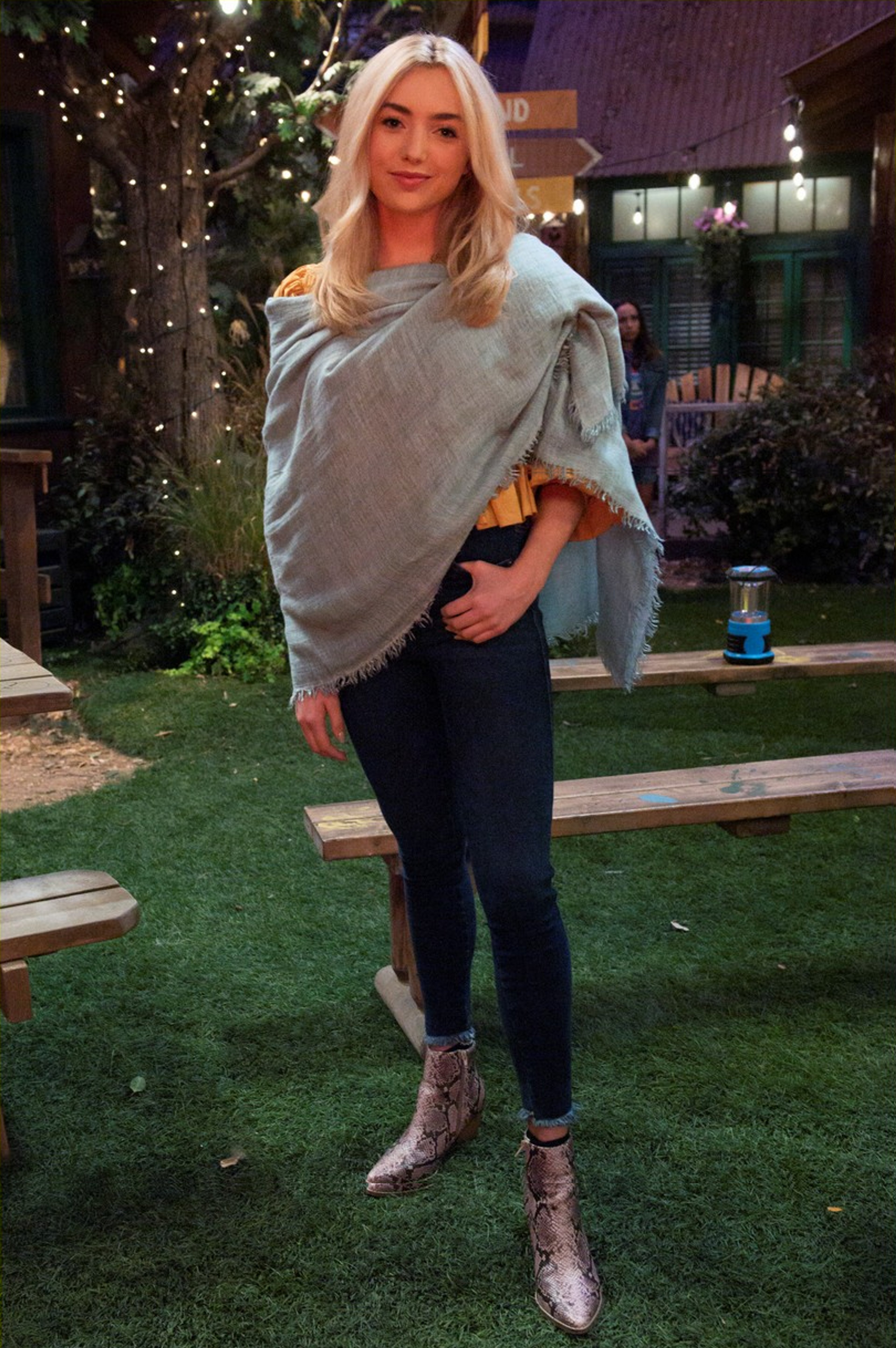 A young woman with long blonde hair wearing a gray sweater standing in front of a festive outdoor setting with Christmas lights and greenery.