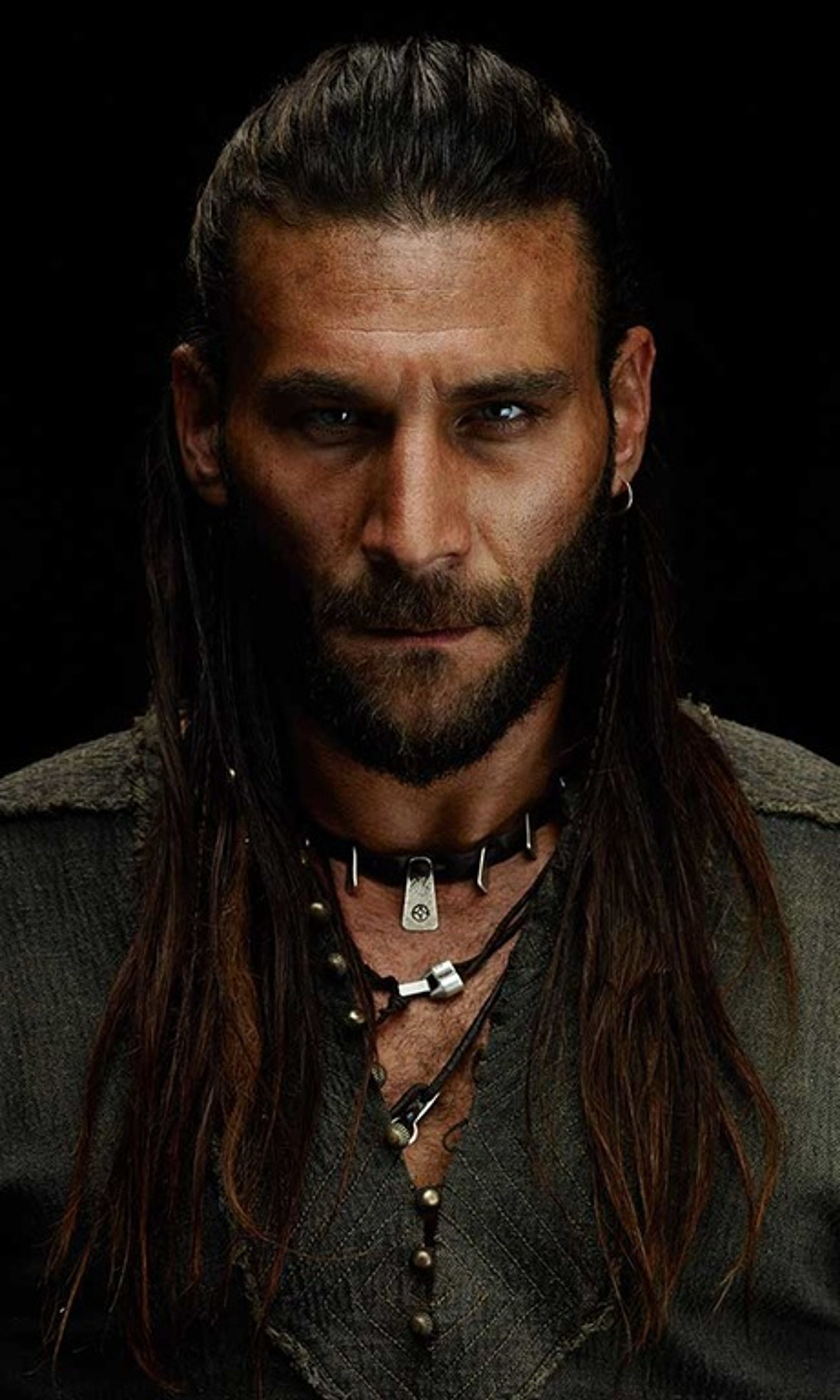 A rugged, bearded man with a piercing gaze, wearing a dark outfit with accessories.