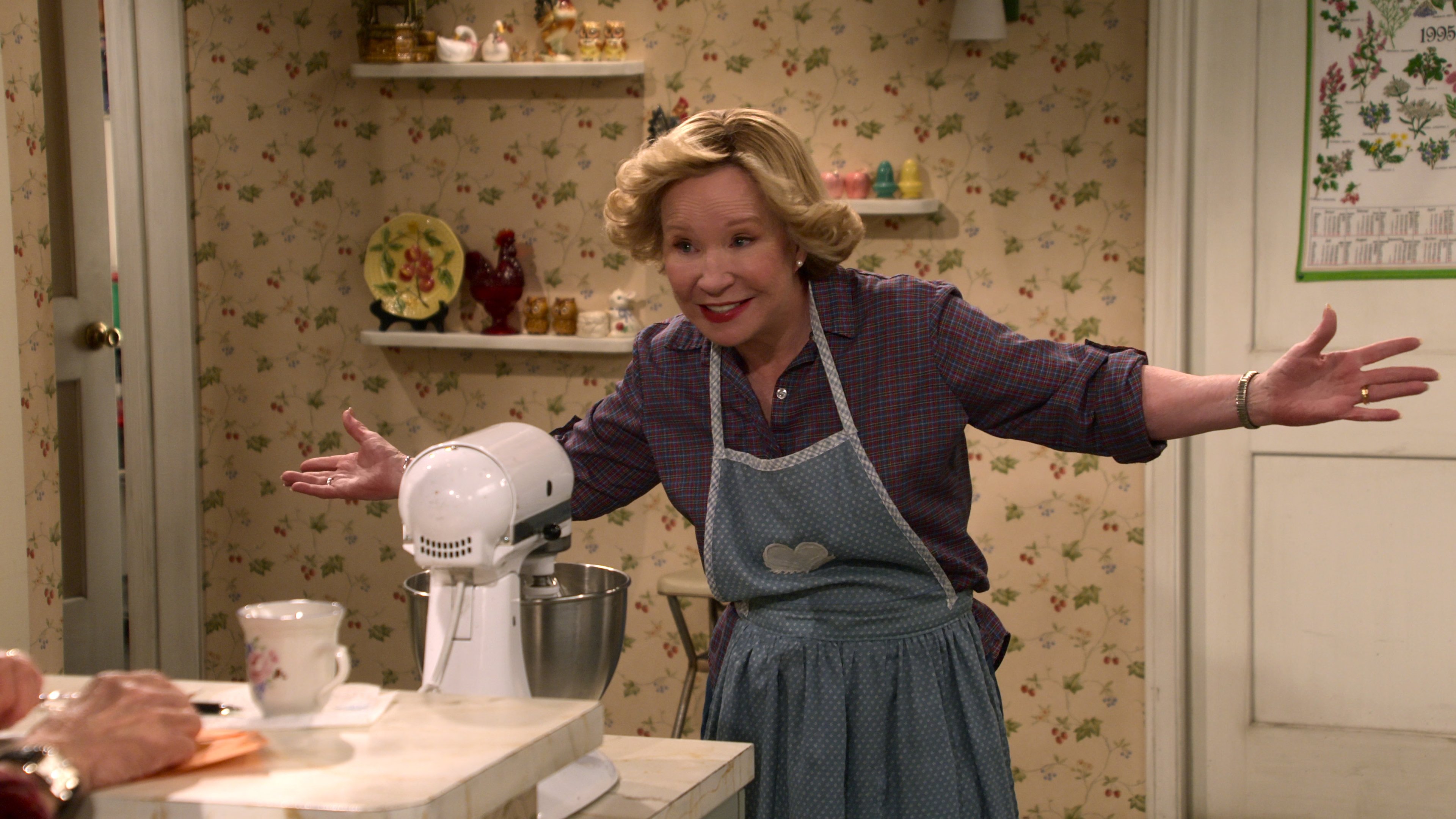 A middle-aged woman in a kitchen setting, gesturing with her hands and smiling.