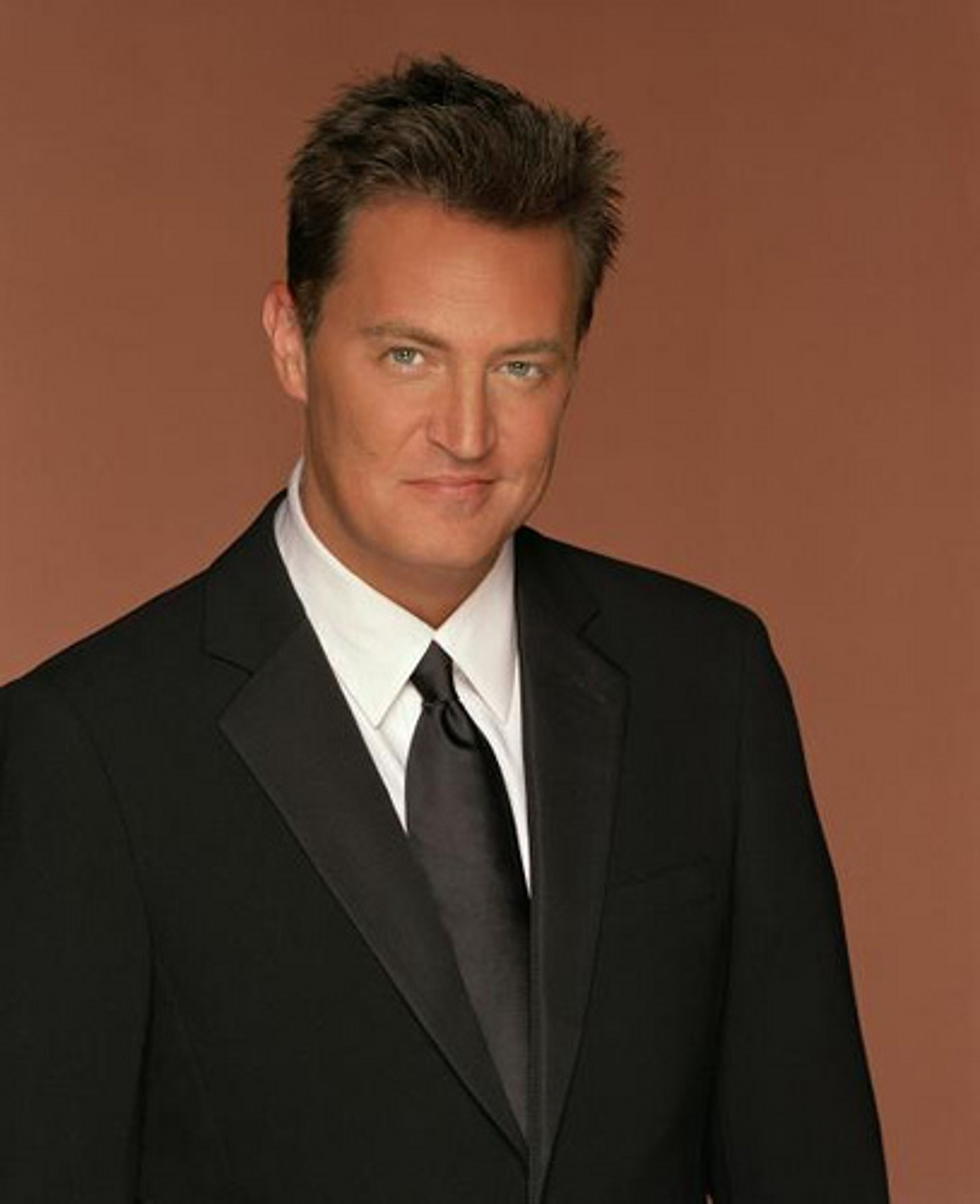 A man in a black suit and tie standing against a brown background