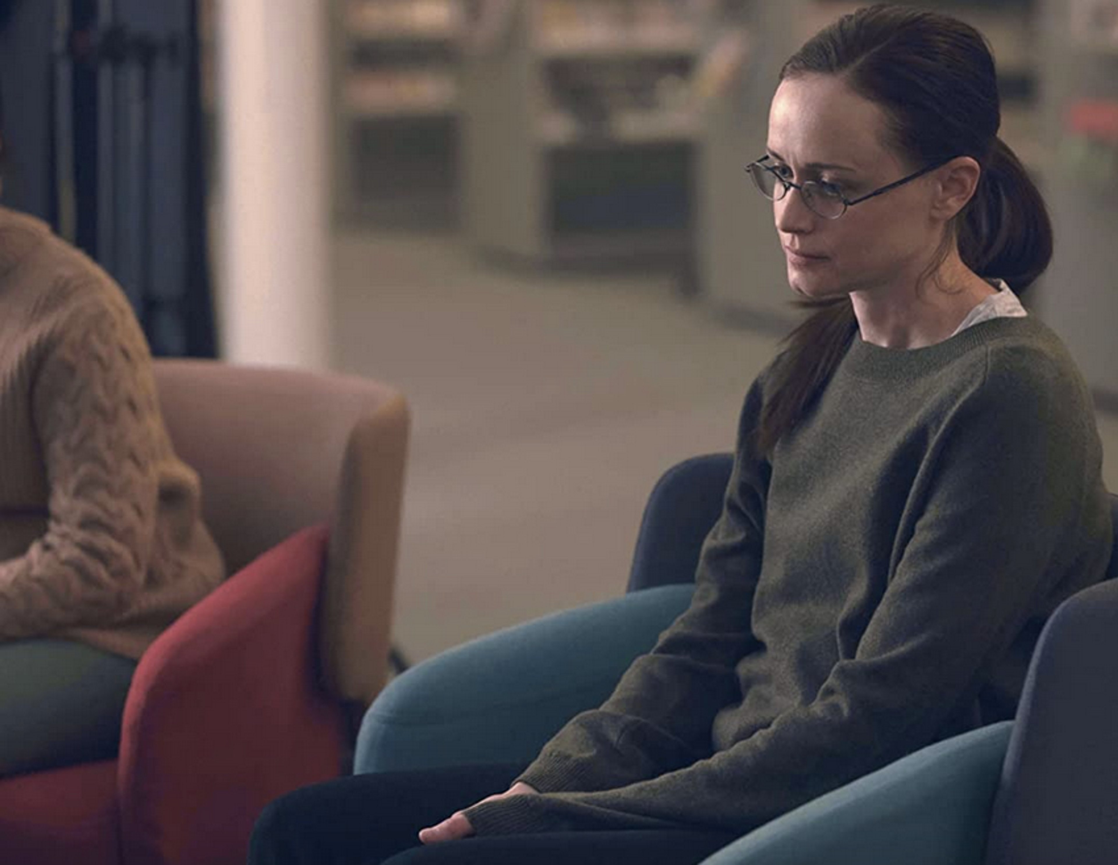 A woman with dark hair and glasses sitting on a couch or chair in a living room setting.