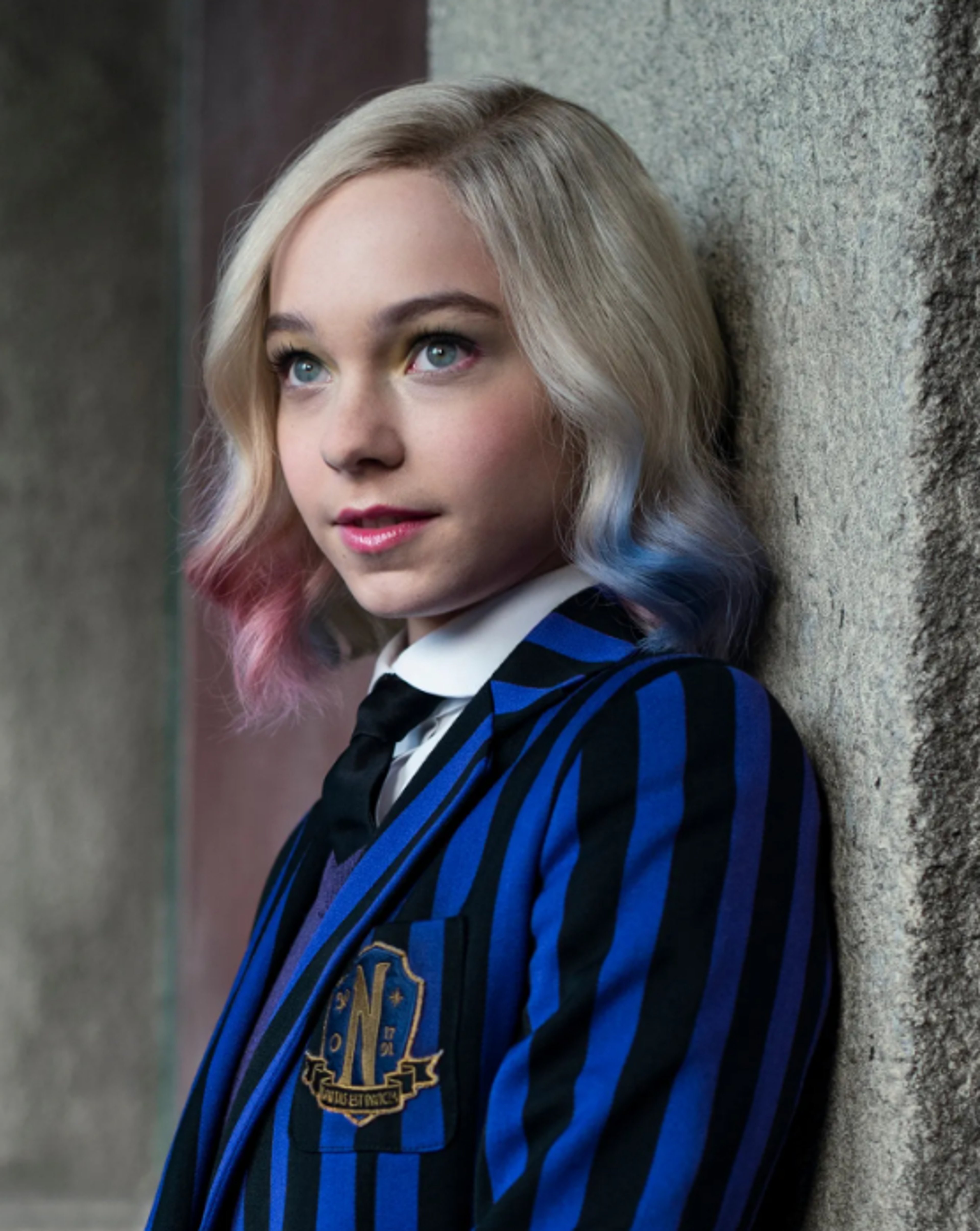 A young woman with blonde and pink hair wearing a striped school uniform, leaning against a wall and smiling.